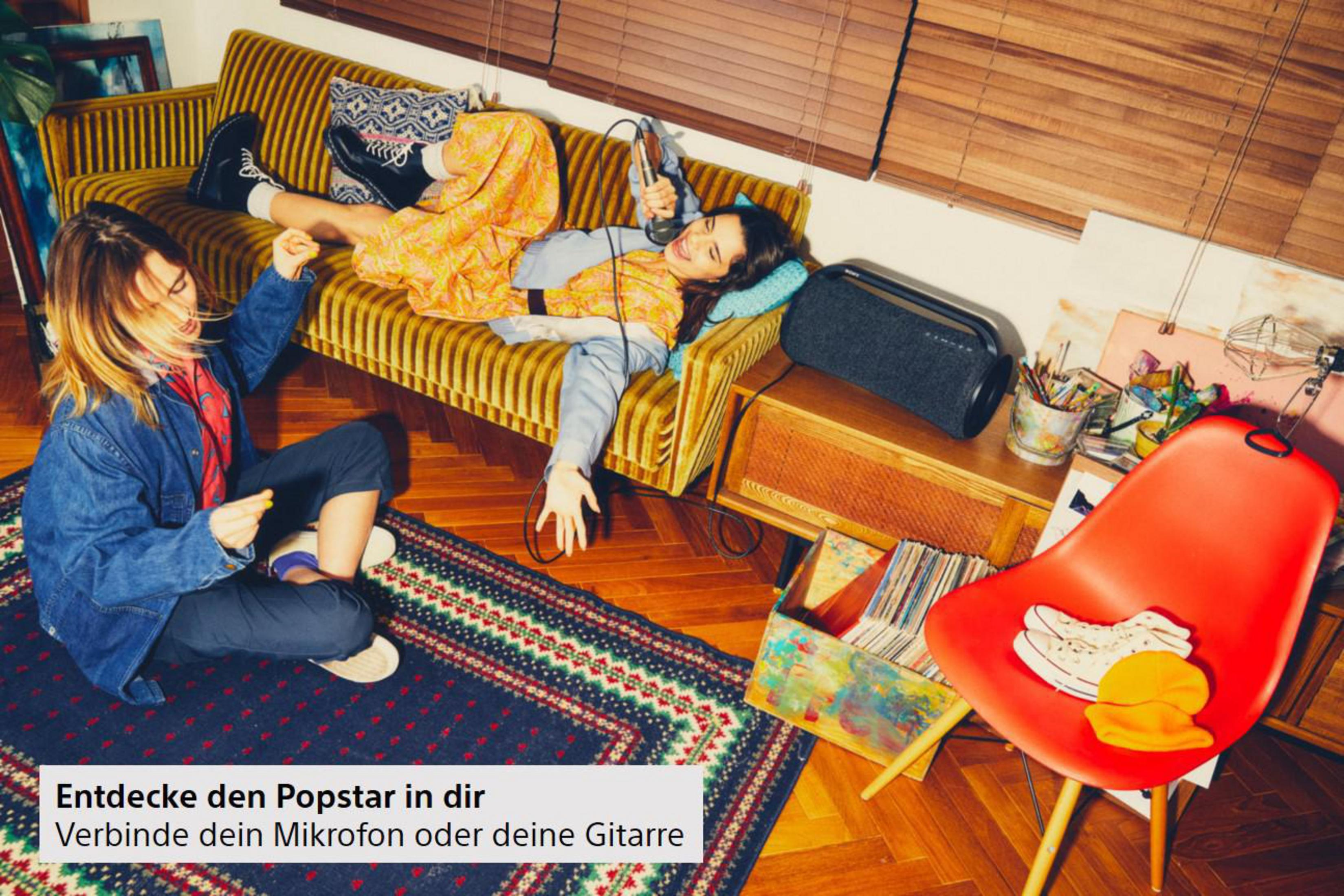 Zwei Personen auf einer Couch mit Lautsprecher. Einer singt ins Mikrofon. Bunte Szene.