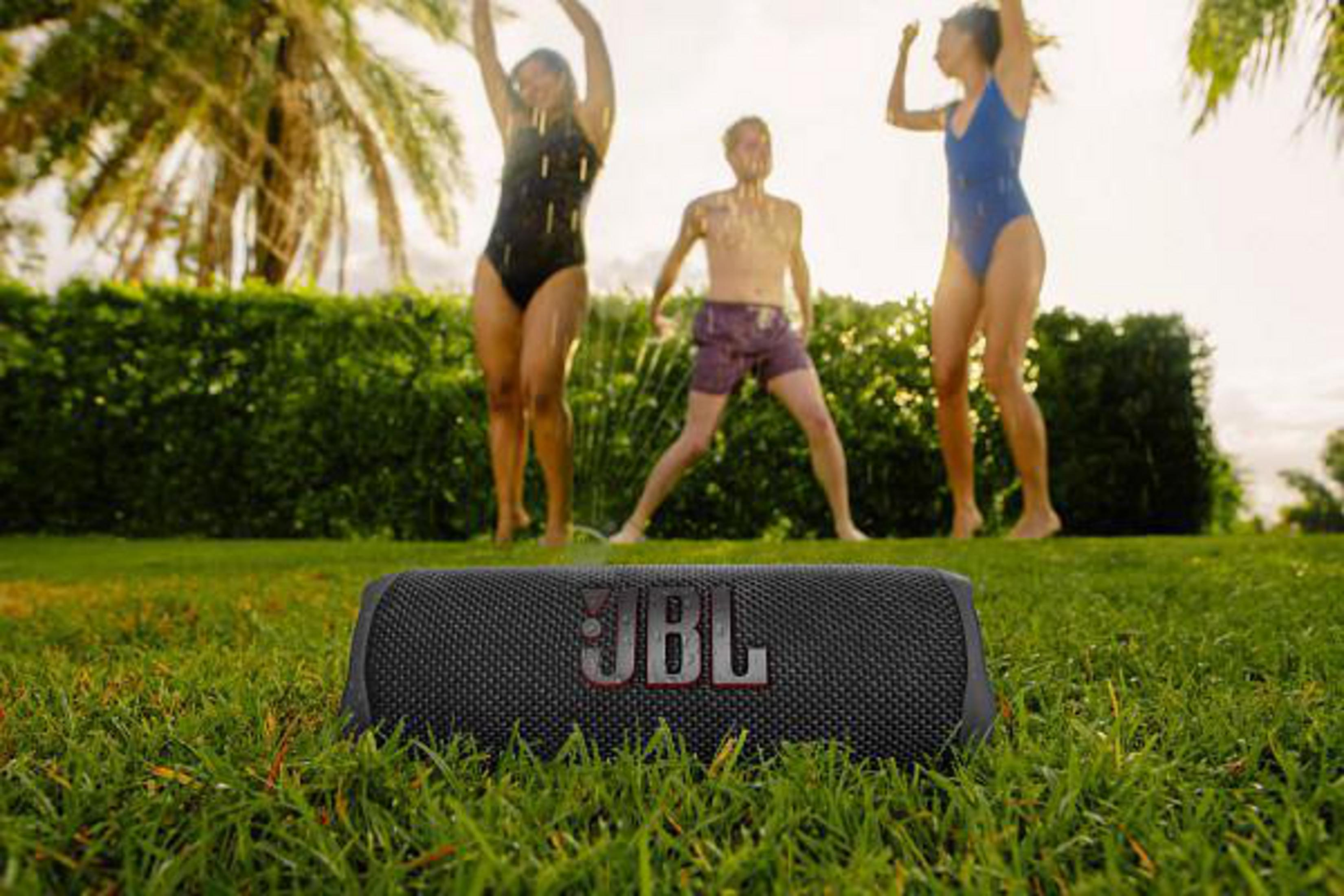 Altavoz JBL en la hierba. Personas al fondo, agua rociando. Escena soleada, al aire libre.