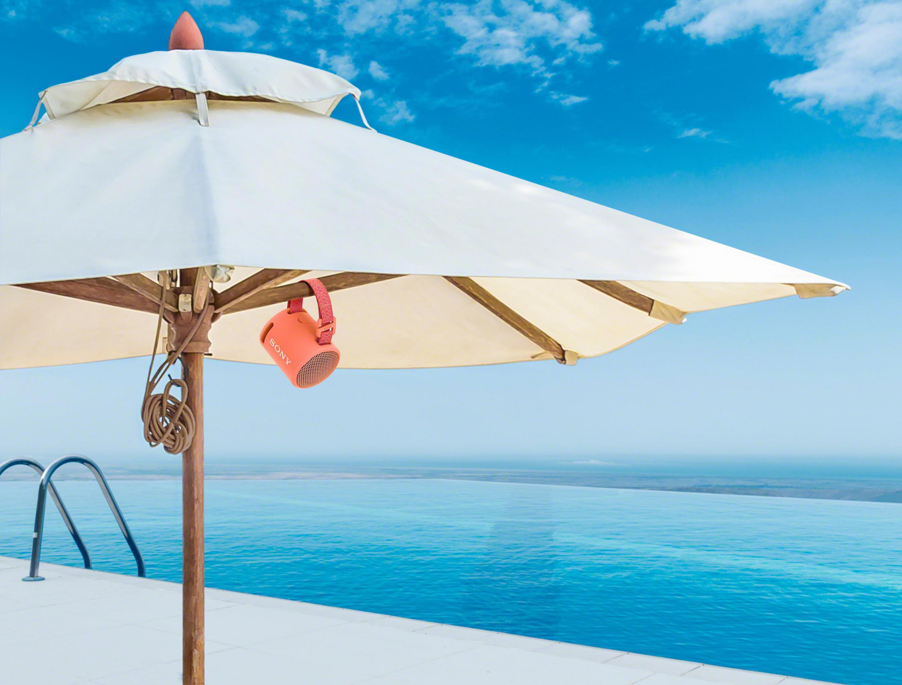 Pomarańczowy głośnik wiszący pod białym parasolem w pobliżu basenu bez krawędzi i oceanu.