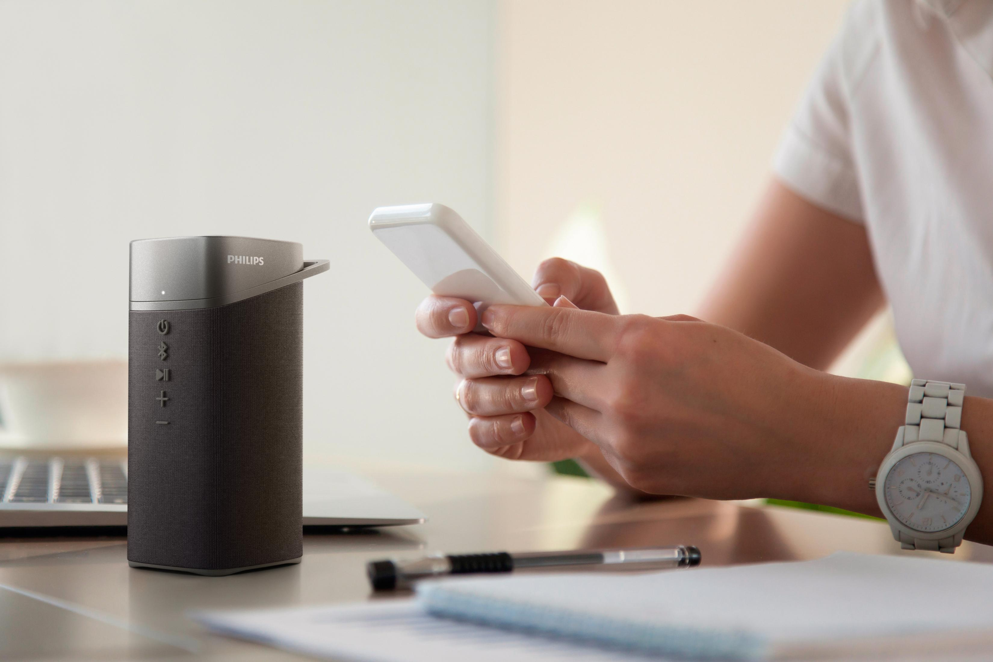 Una persona tiene in mano uno smartphone bianco, con un altoparlante e oggetti da scrivania nella scena.