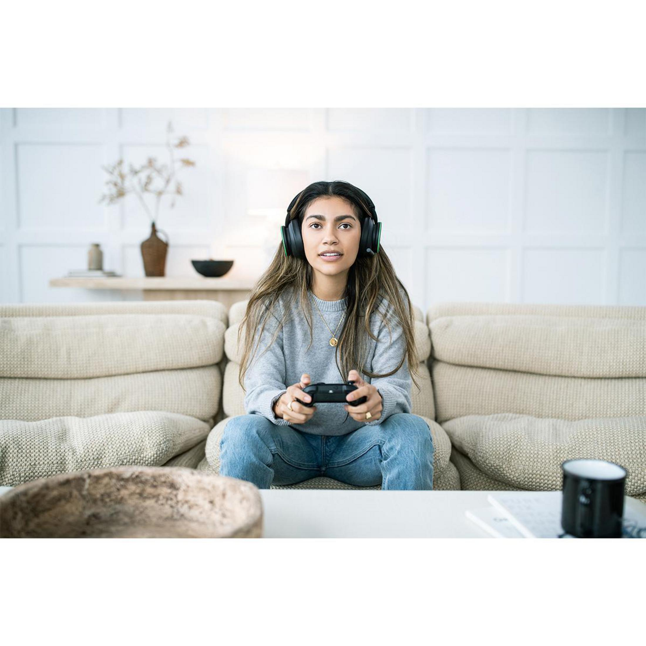 Kobieta gra w grę wideo na kanapie, trzyma kontroler, ma na sobie słuchawki.