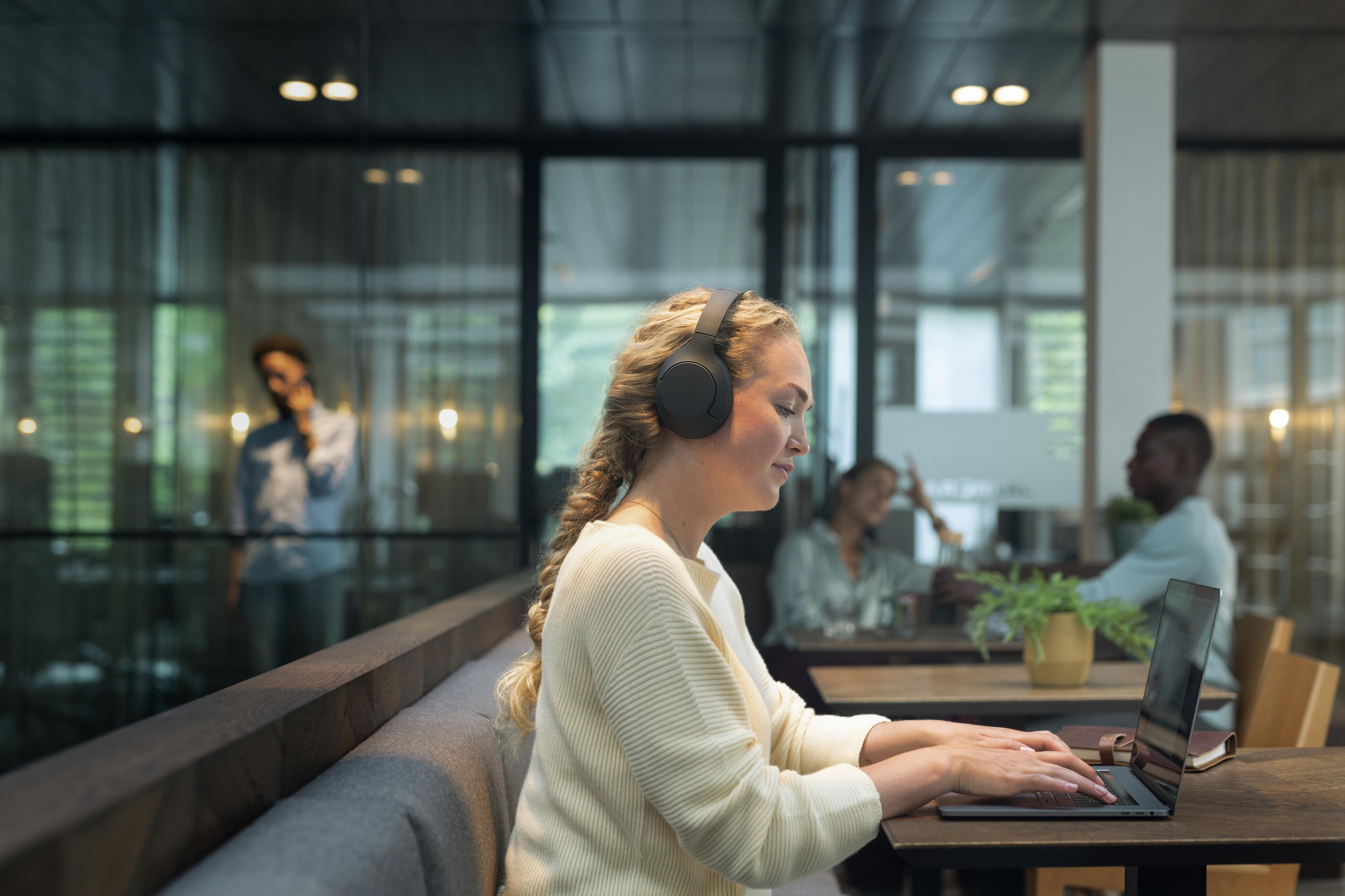 Eine Frau mit blonden Haaren, die Kopfhörer trägt, tippt in einem Café auf einem Laptop. Andere Leute im Hintergrund.