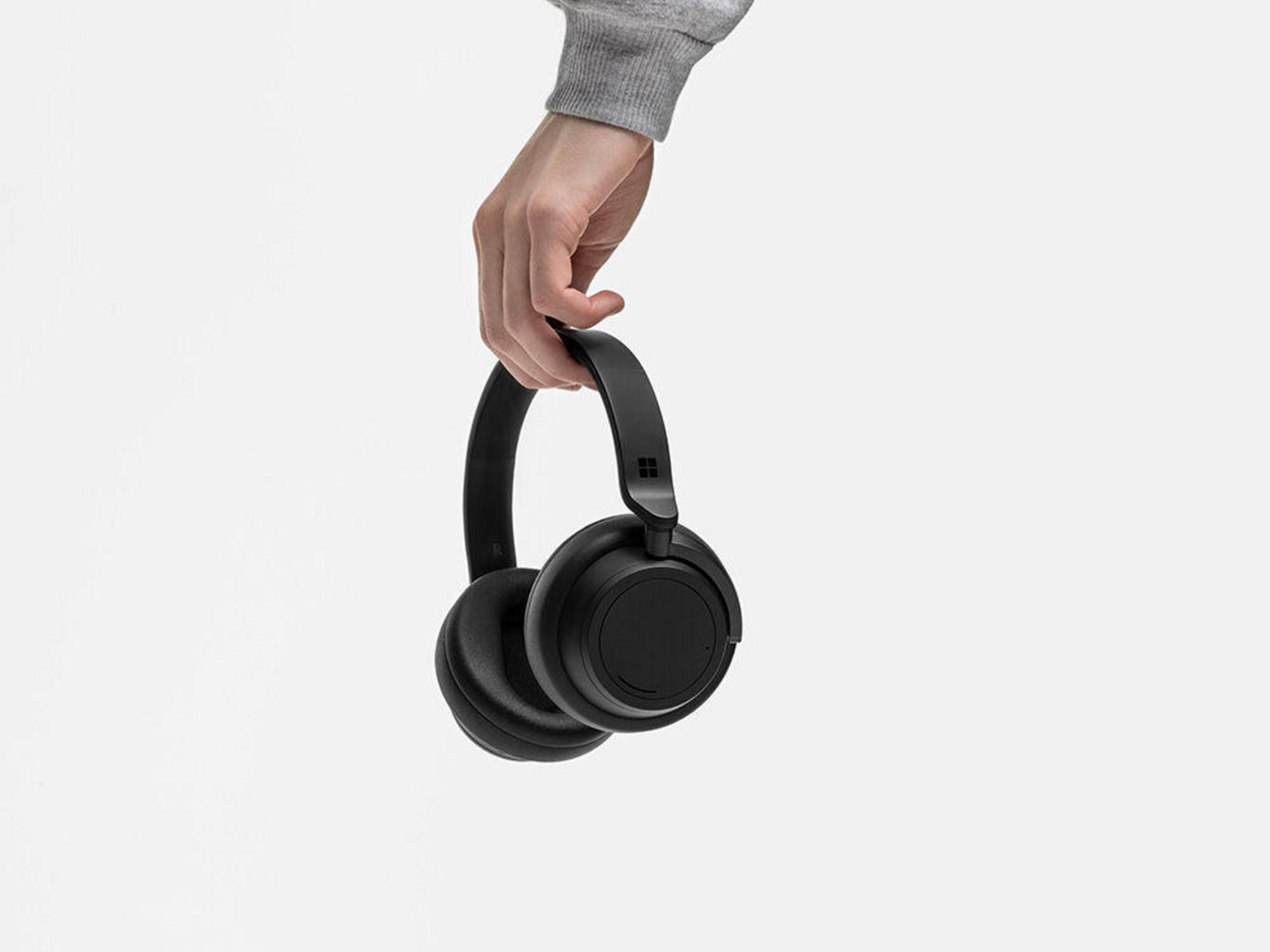 Una mano che tiene un paio di cuffie Microsoft Surface nere contro uno sfondo bianco.