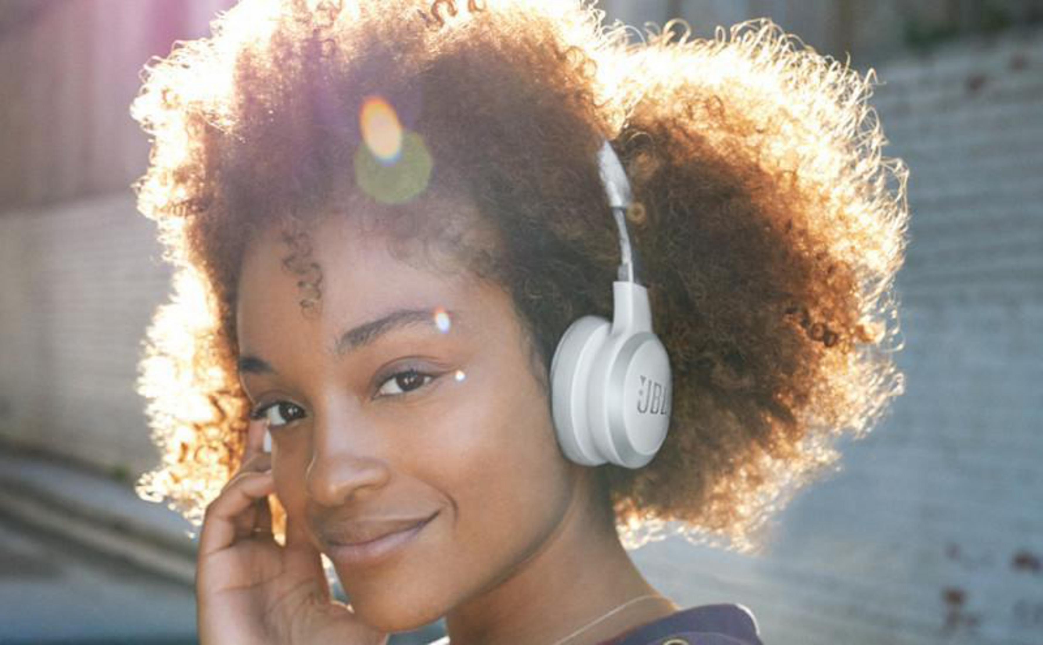 Mujer con pelo rizado con auriculares JBL blancos, sol brilla. Sonríe, se agarra la oreja.