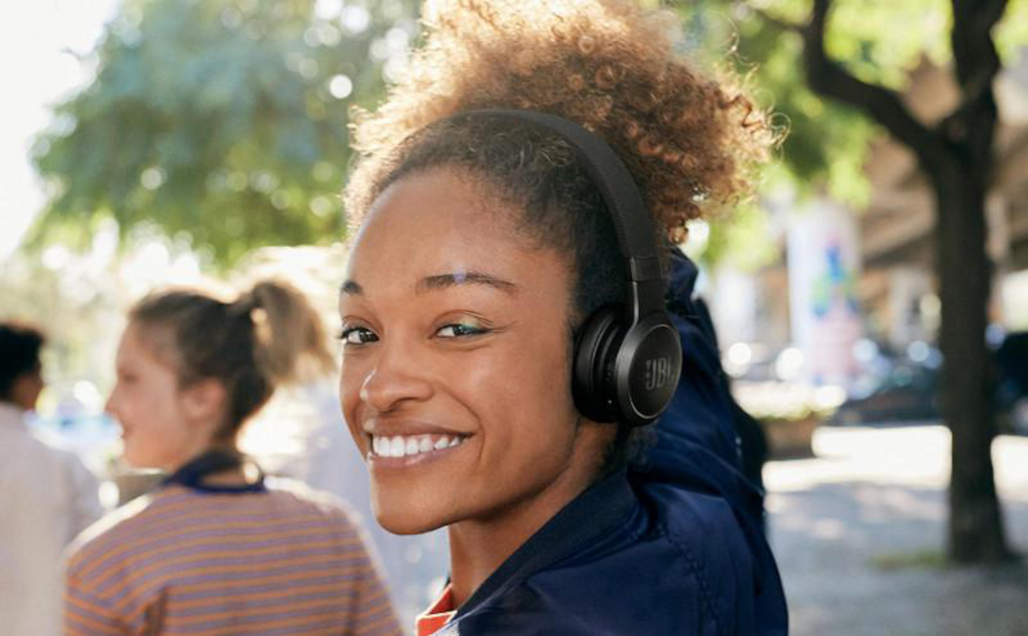 Mujer sonriendo con auriculares JBL negros caminando en la calle, fondo personas.