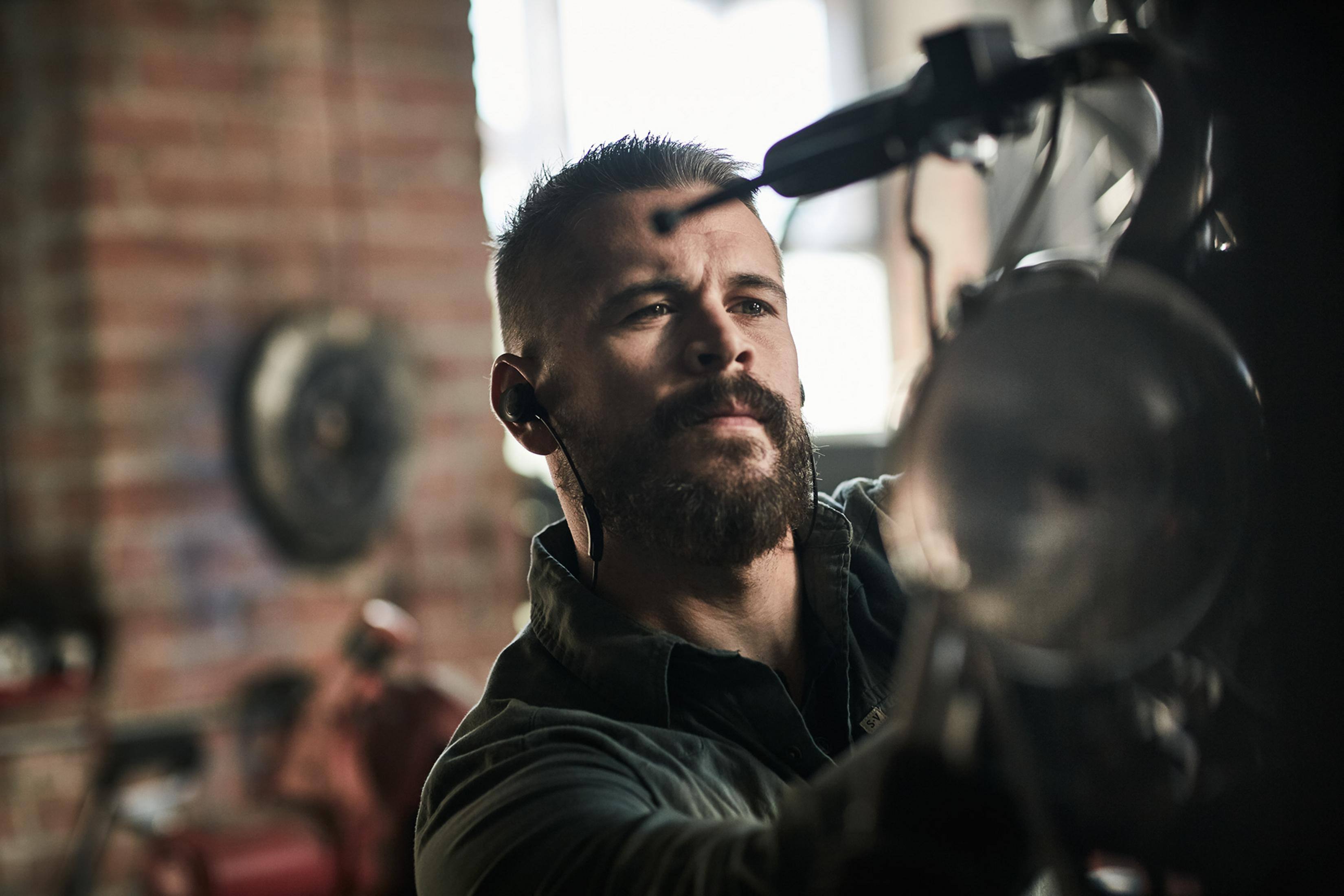 Un hombre con barba que usa auriculares está trabajando en una motocicleta en un taller.