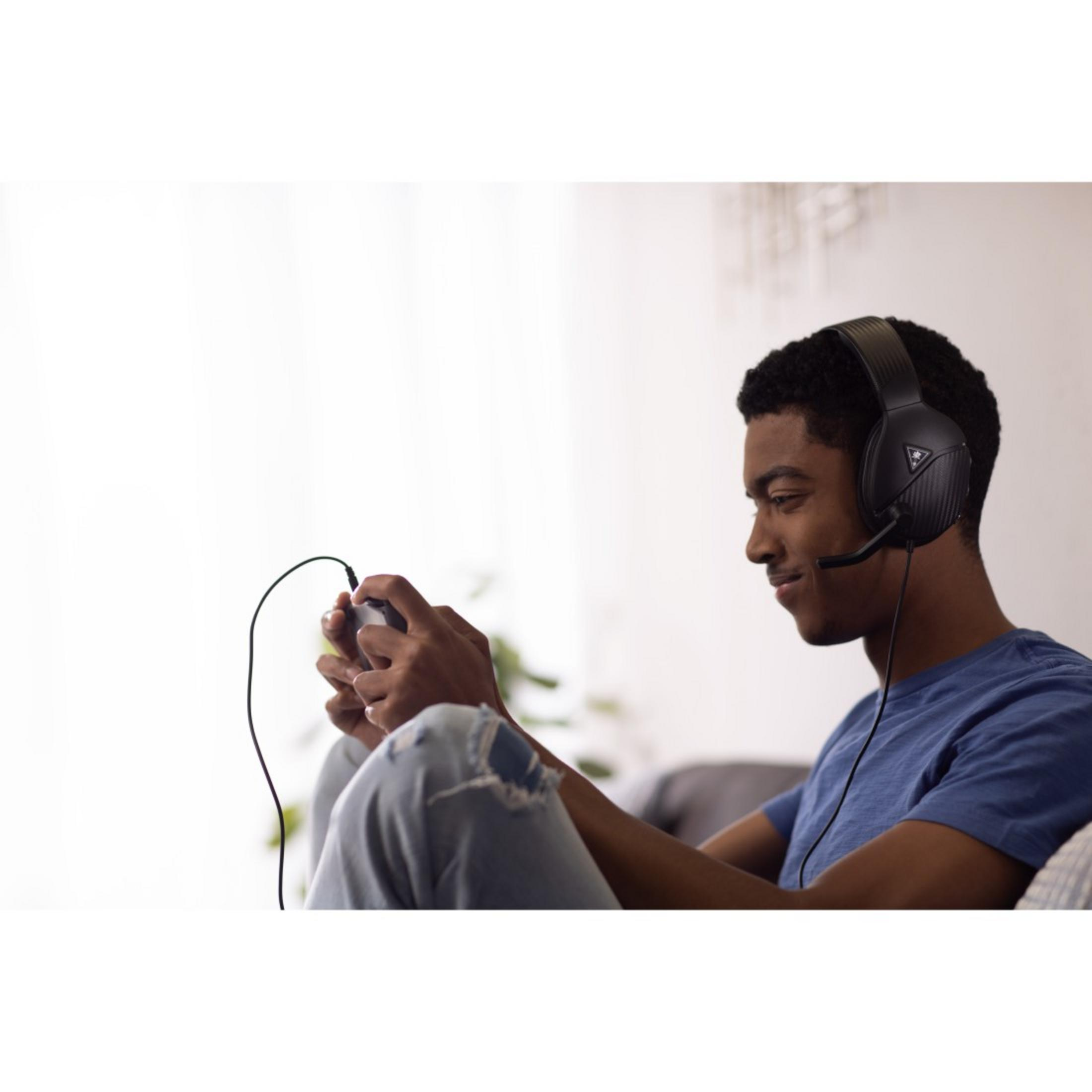 Młody mężczyzna grający w grę, w zestawie słuchawkowym z mikrofonem, uśmiechnięty, trzymający kontroler.