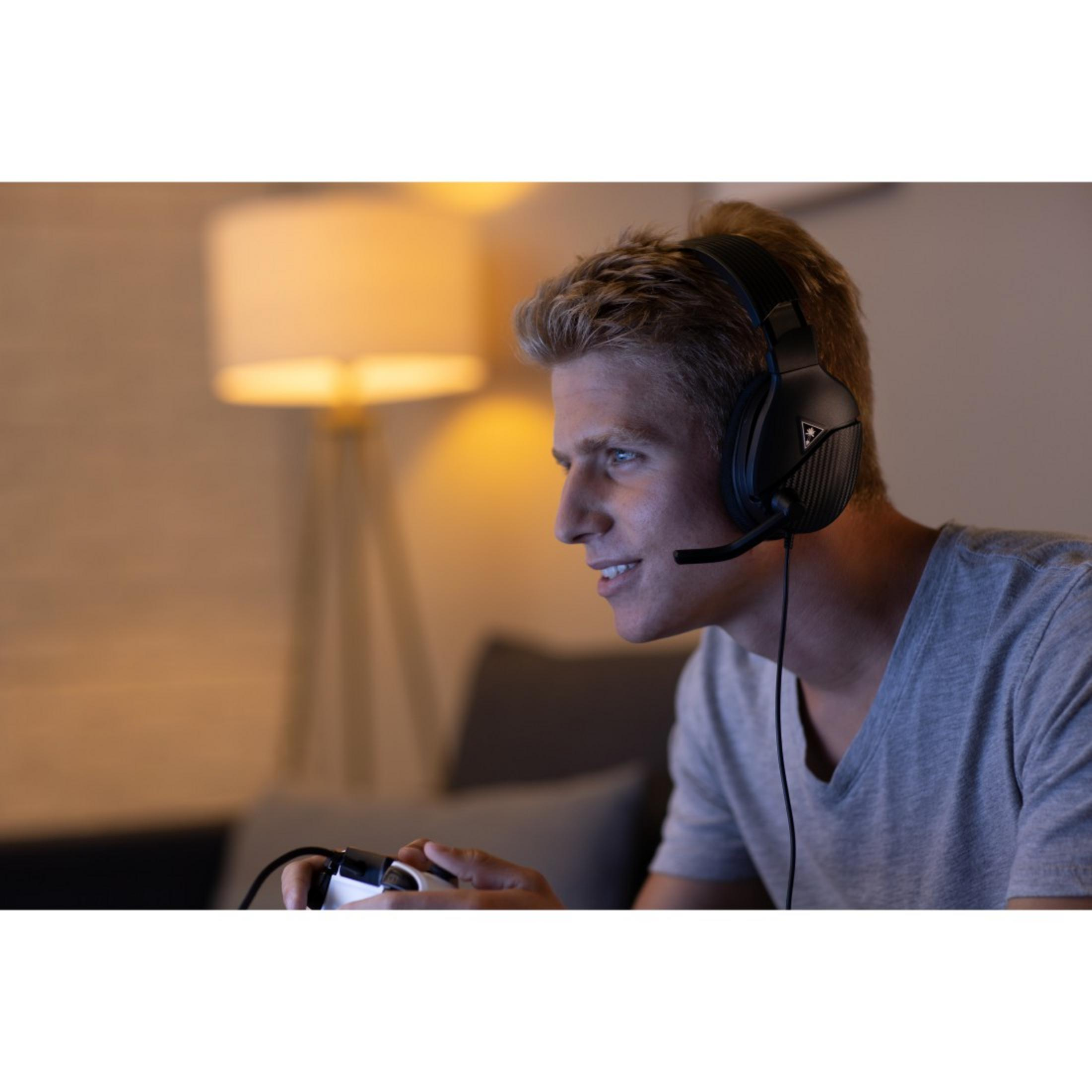 Blondyn w słuchawkach, trzymający kontroler, grający w grę, na tle ciemnego pokoju i lampy.