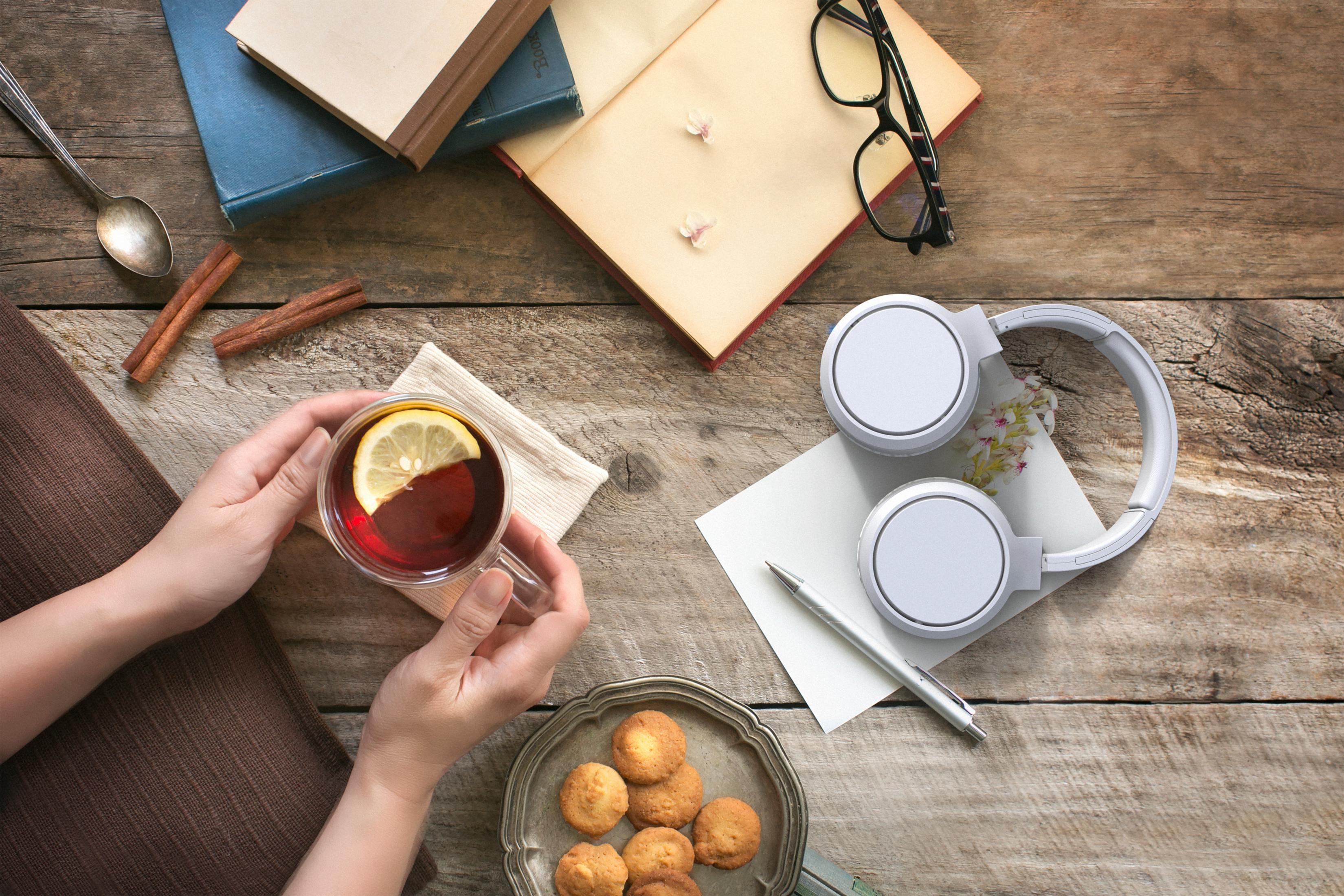 Hände halten Teetasse mit Zitrone, Keksen, Kopfhörern, Büchern und Brille auf Holztisch.