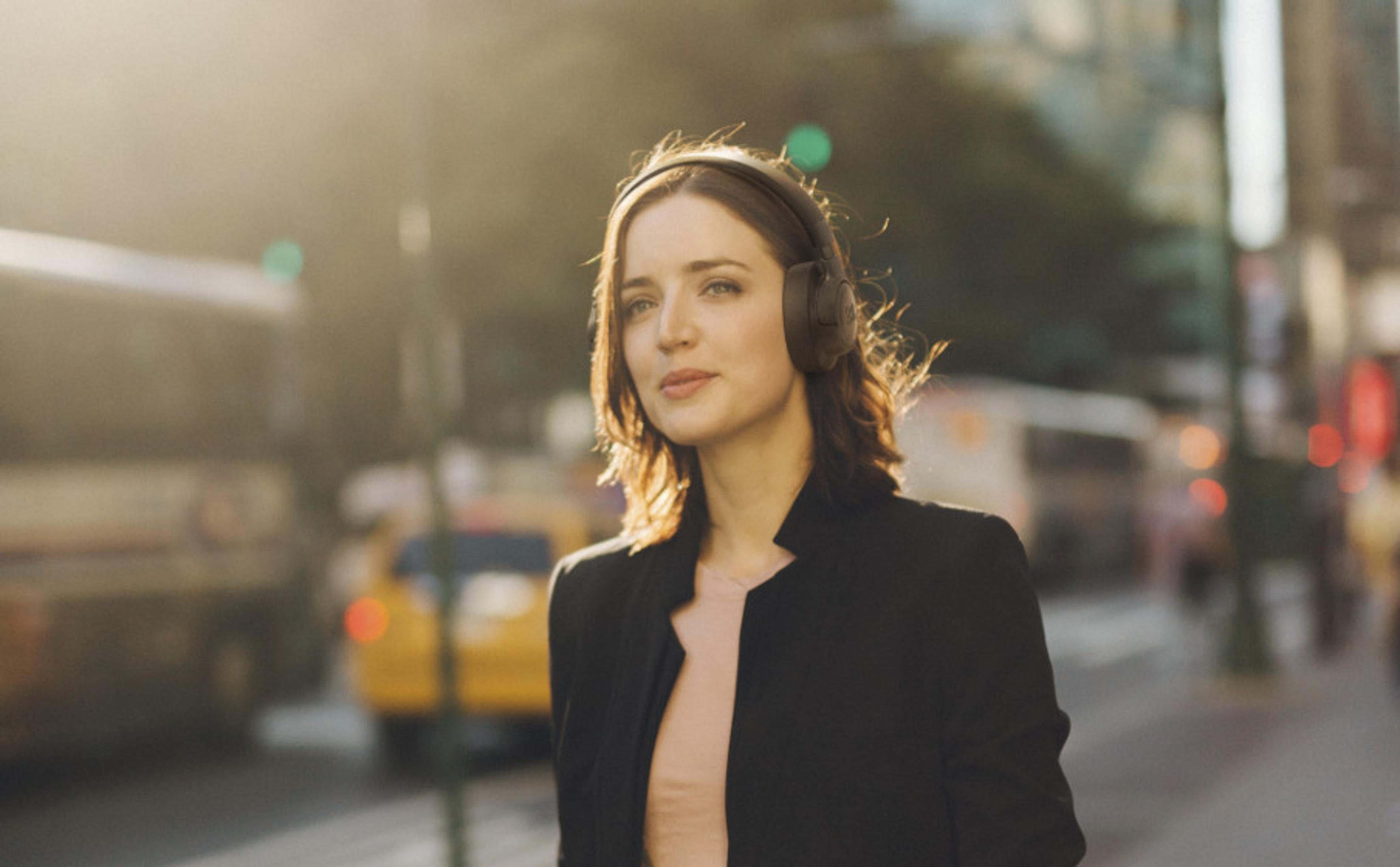 Donna con blazer nero e cuffie che cammina per una strada cittadina, luce solare sullo sfondo.