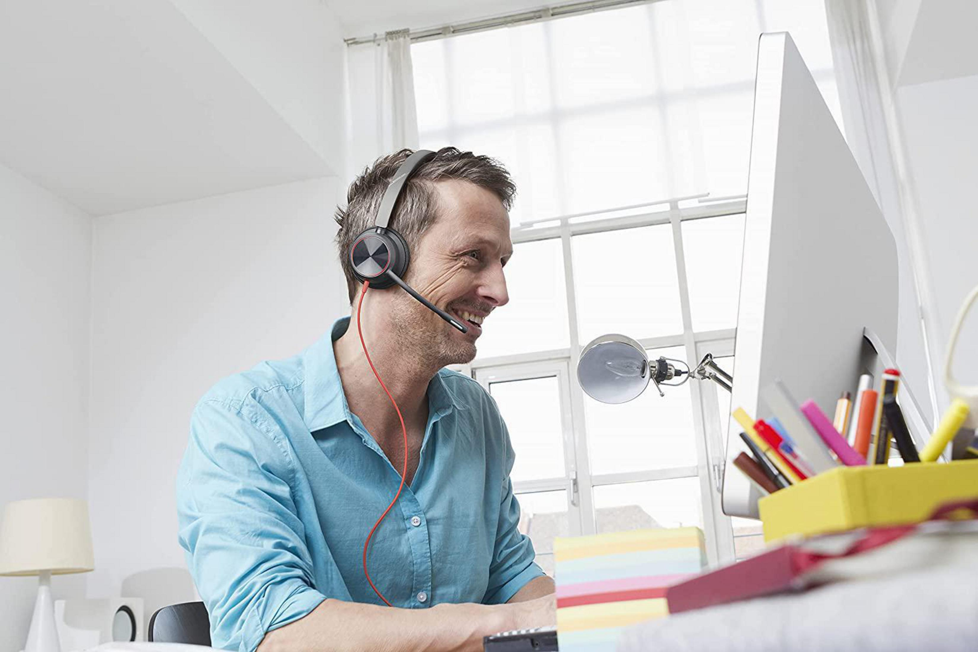 Mężczyzna w słuchawkach uśmiecha się do komputera. Na biurku znajdują się artykuły biurowe. Tłem jest okno.