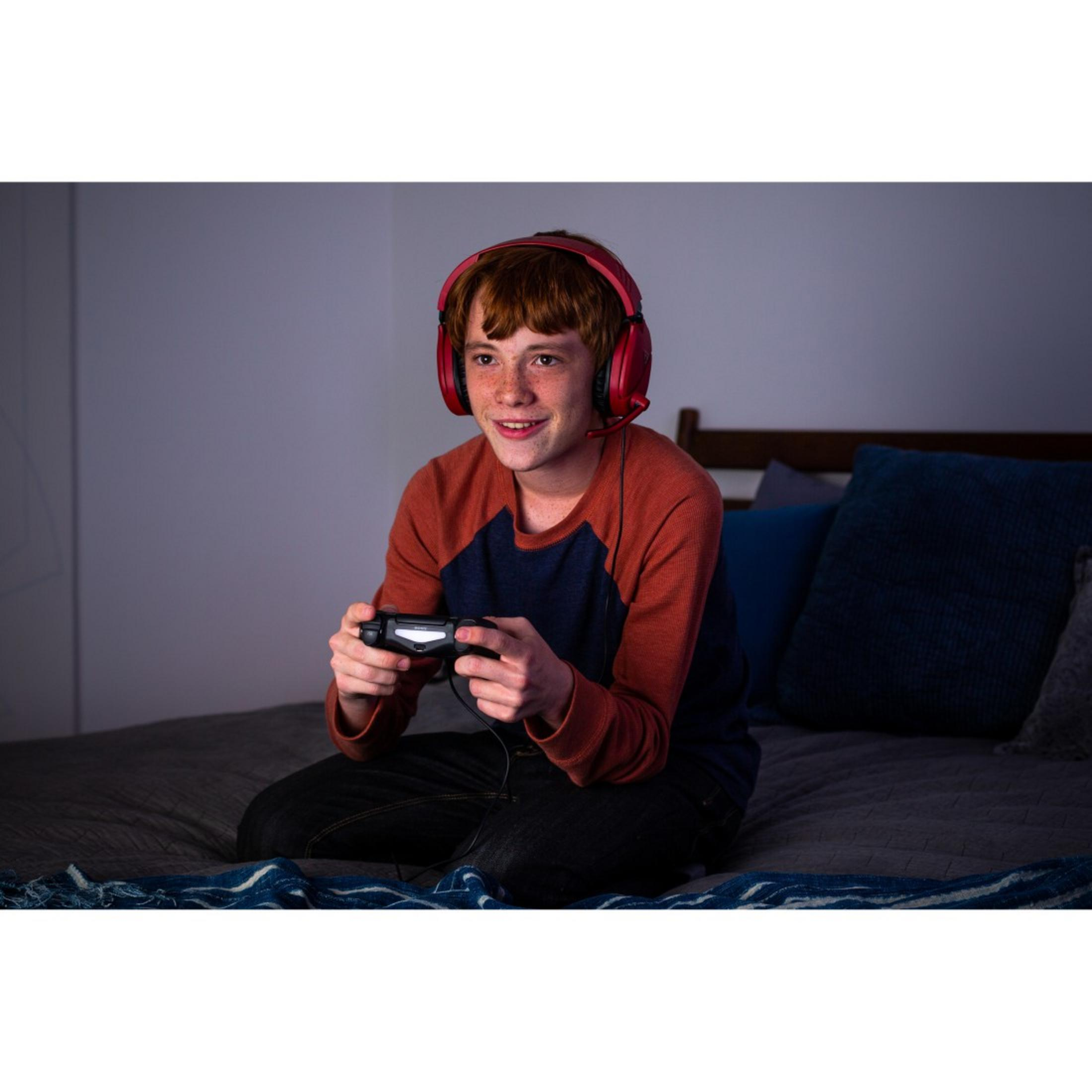 Młody gracz z czerwonym zestawem słuchawkowym gra na łóżku. Trzyma kontroler i uśmiecha się.