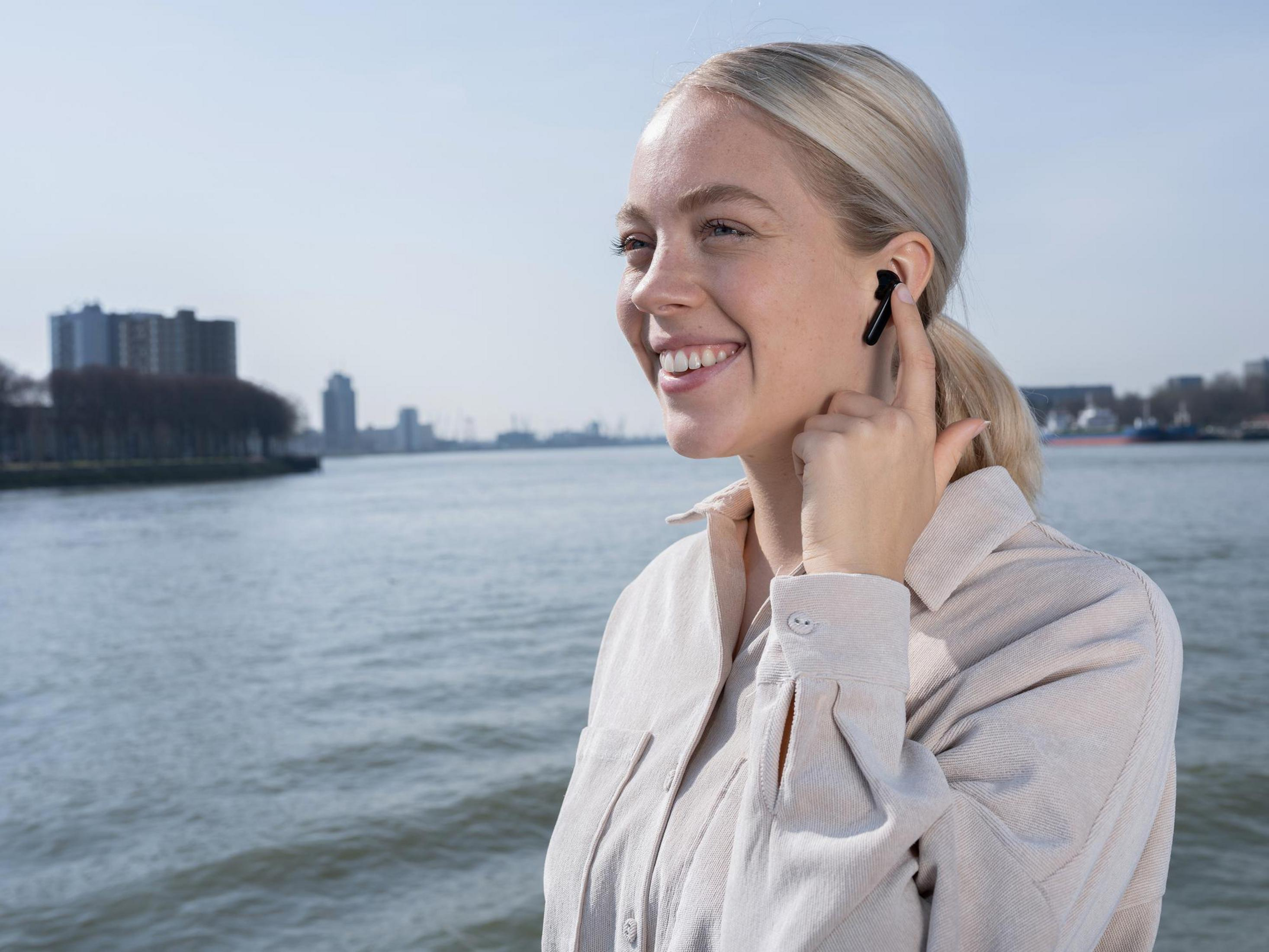 Blondynka z uśmiechem, w beżowej koszuli, trzyma słuchawkę w pobliżu rzeki.