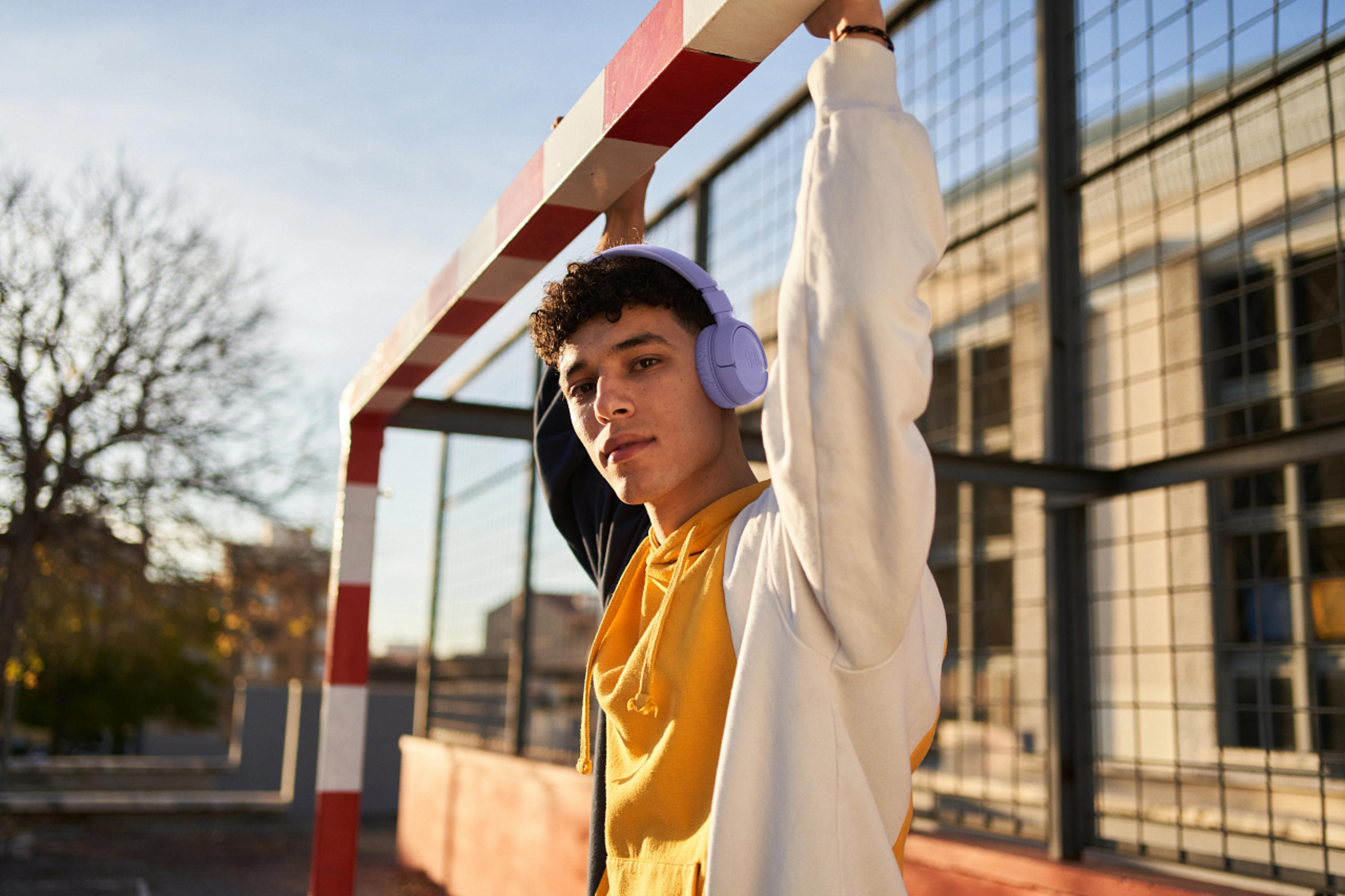 Młody mężczyzna ze słuchawkami stoi przy bramce sportowej. Ma na sobie żółto-białą bluzę z kapturem.