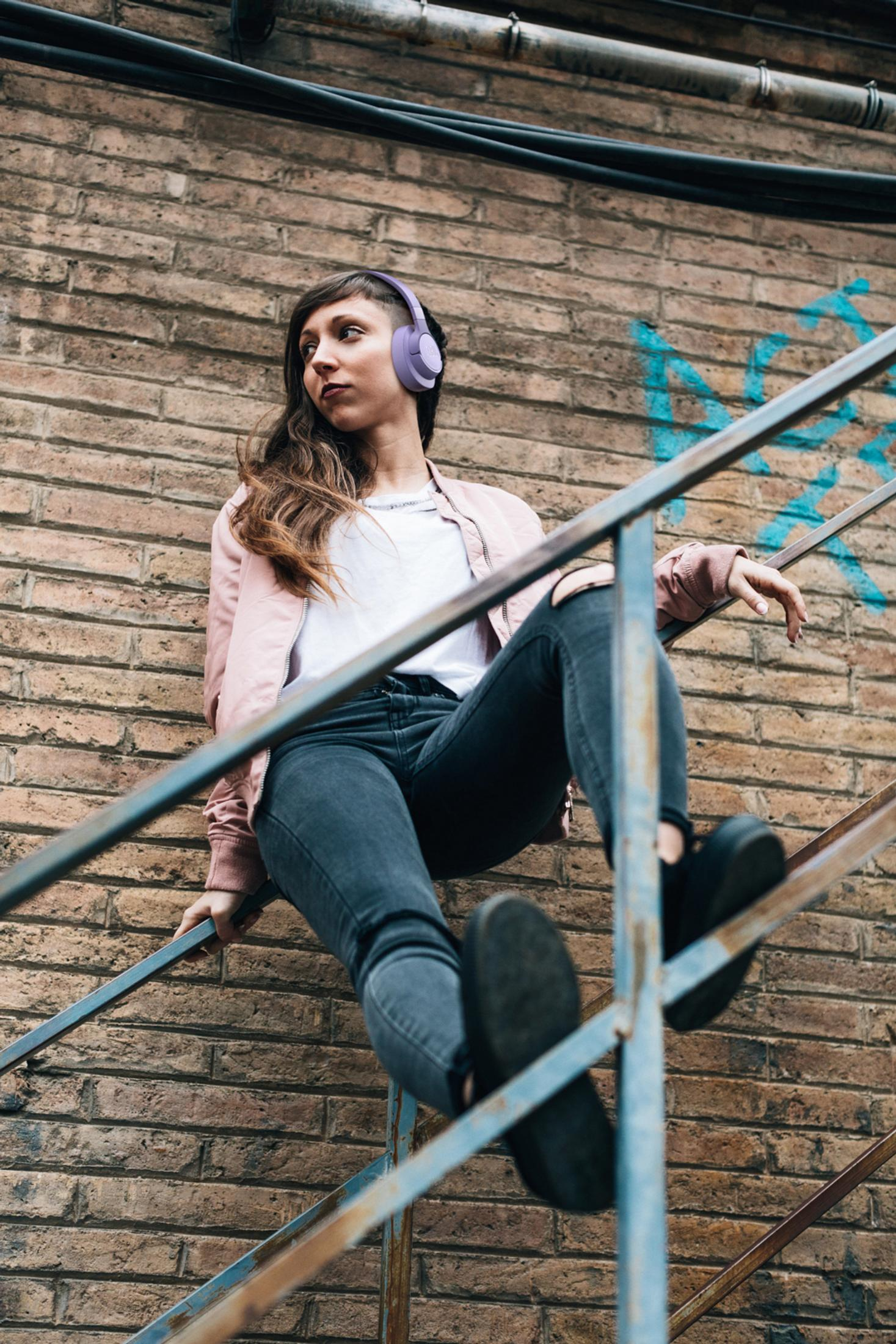 Kobieta siedząca na schodach ze słuchawkami. Ubrana w różową kurtkę i dżinsy. Tło z ceglanego muru.