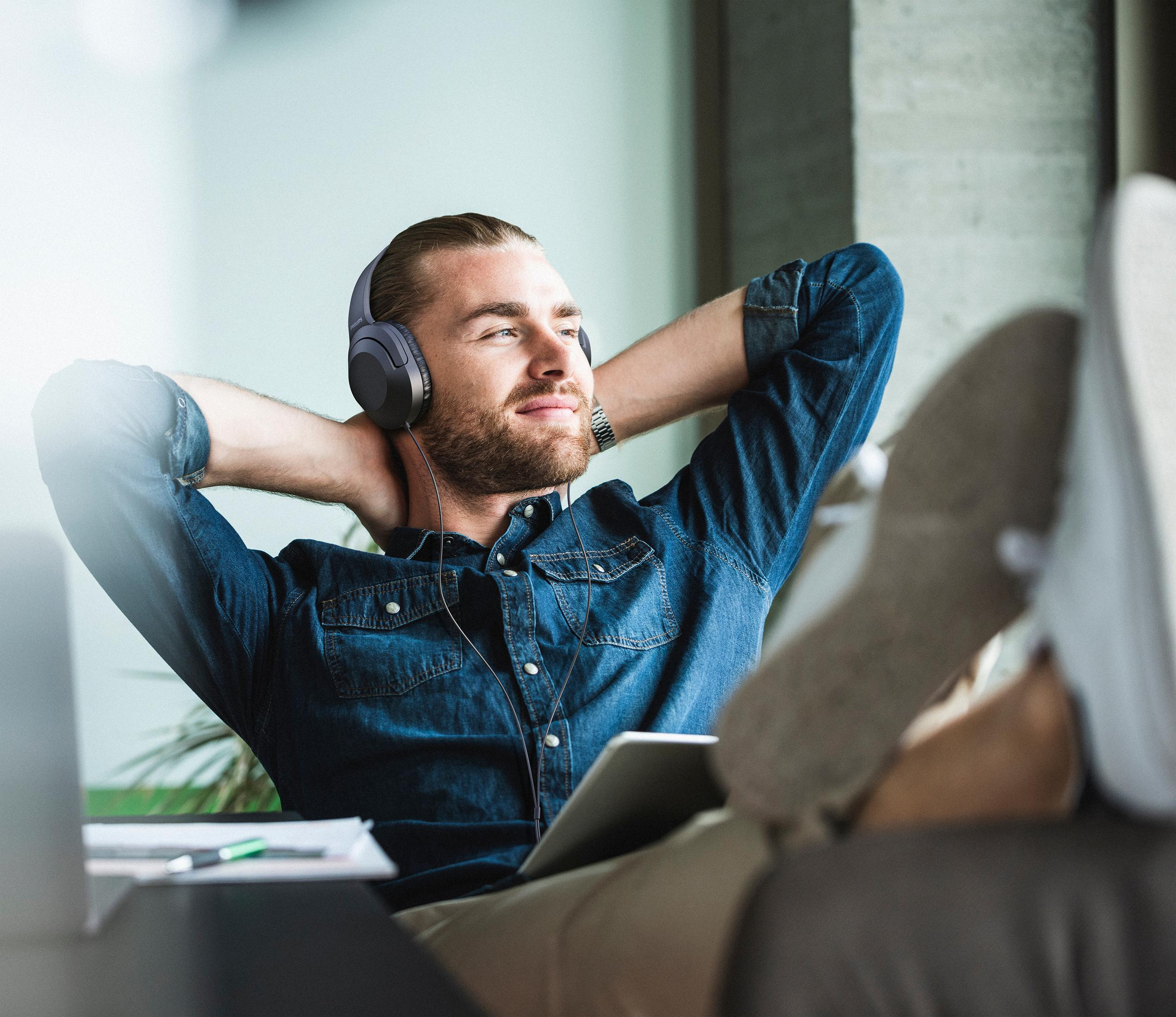 Mężczyzna w słuchawkach, uśmiechnięty, z tabletem na kolanach, relaksujący się w pomieszczeniu.