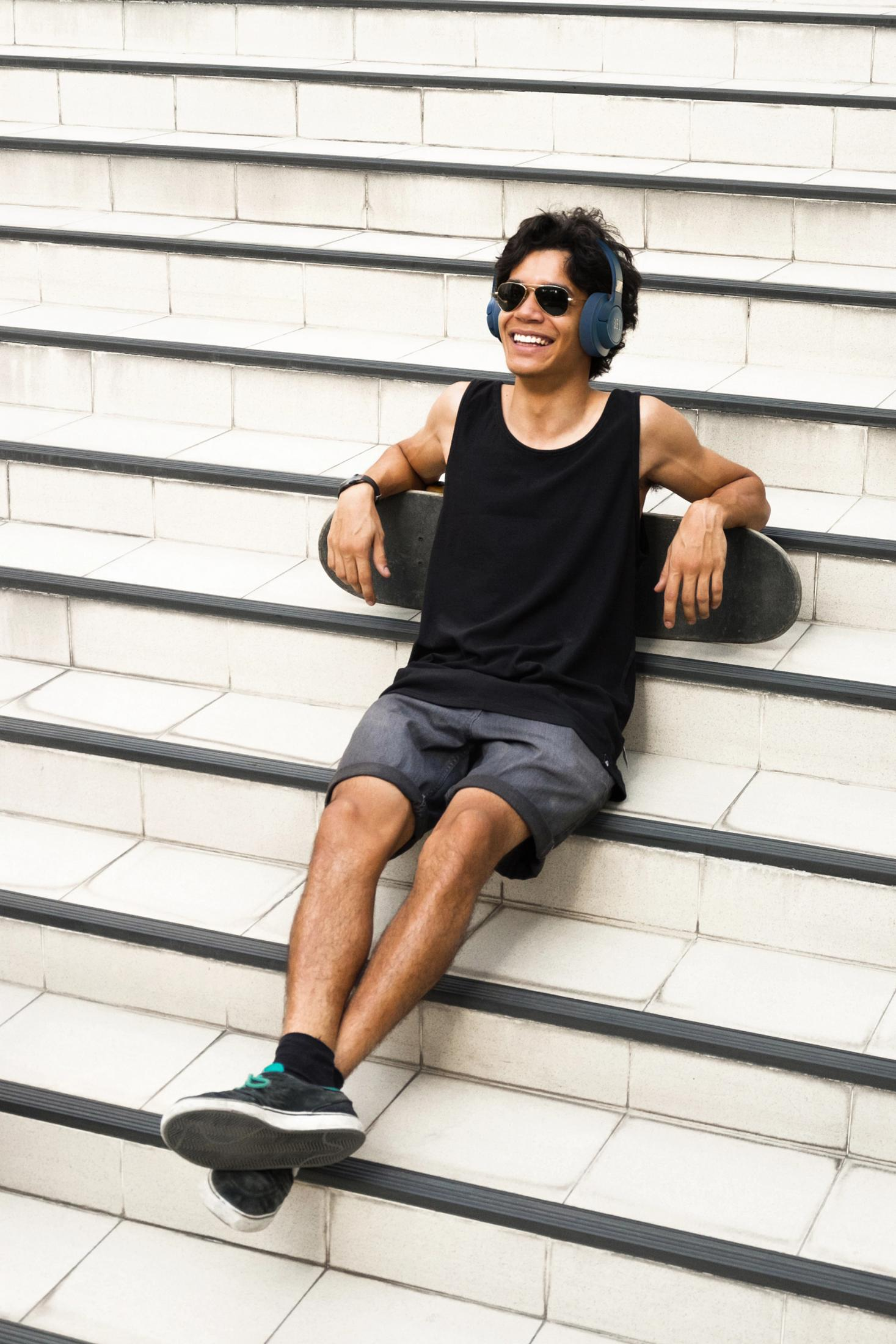 Młody mężczyzna na schodach w okularach przeciwsłonecznych i słuchawkach, trzyma deskorolkę.