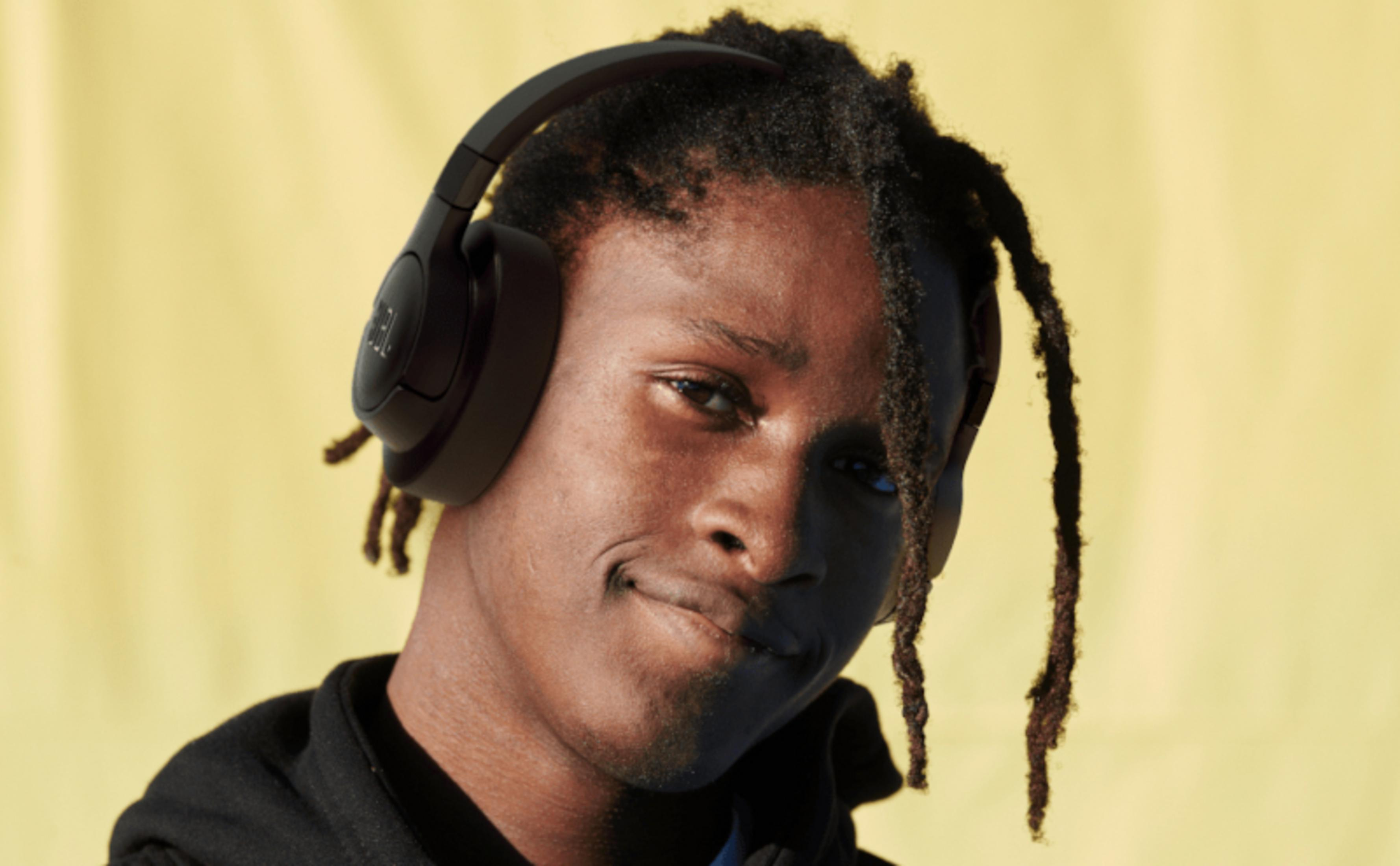 Uomo con dreadlocks con cuffie JBL, sorridente, con sfondo giallo.