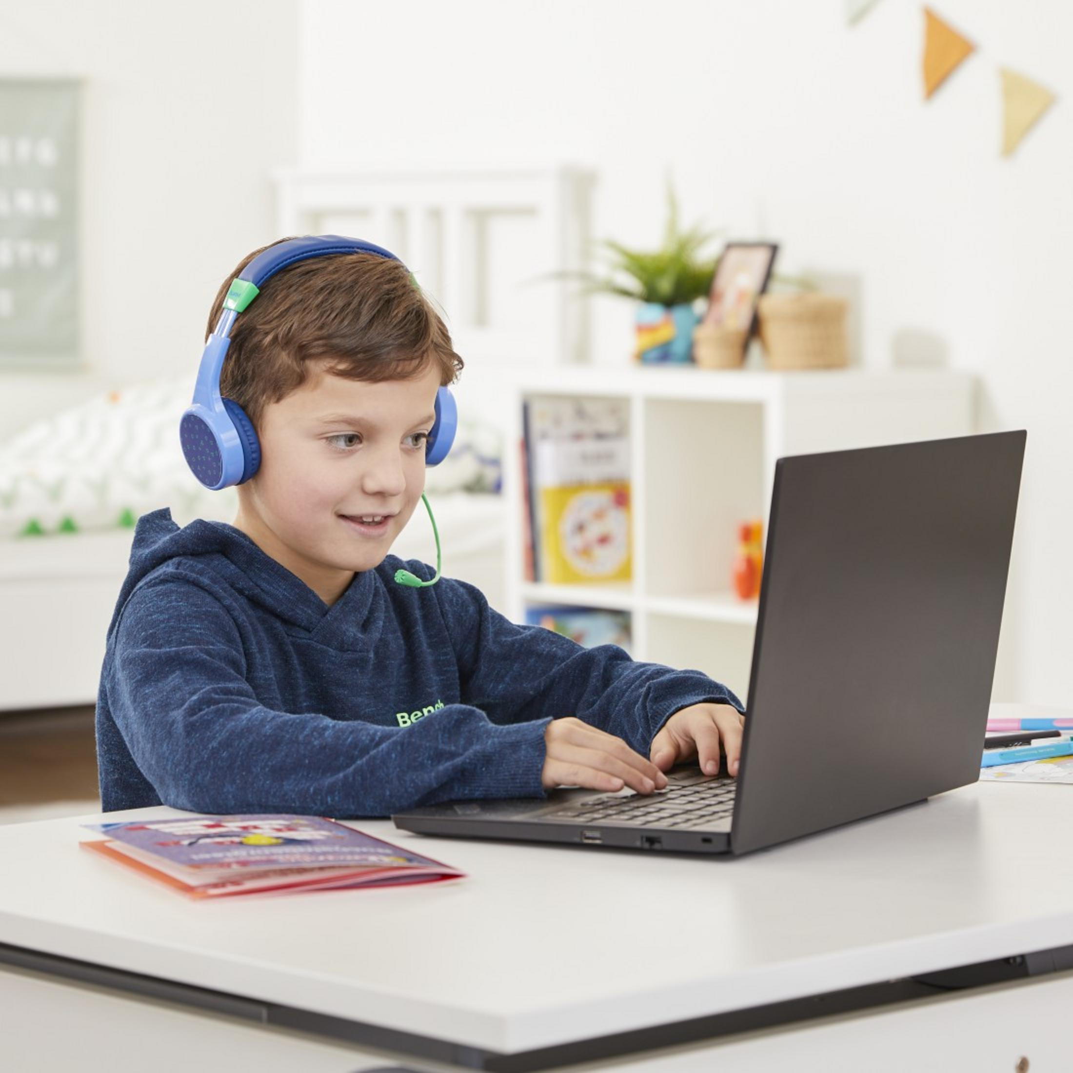 Un ragazzino usa un laptop con cuffie blu e un libro su una scrivania.