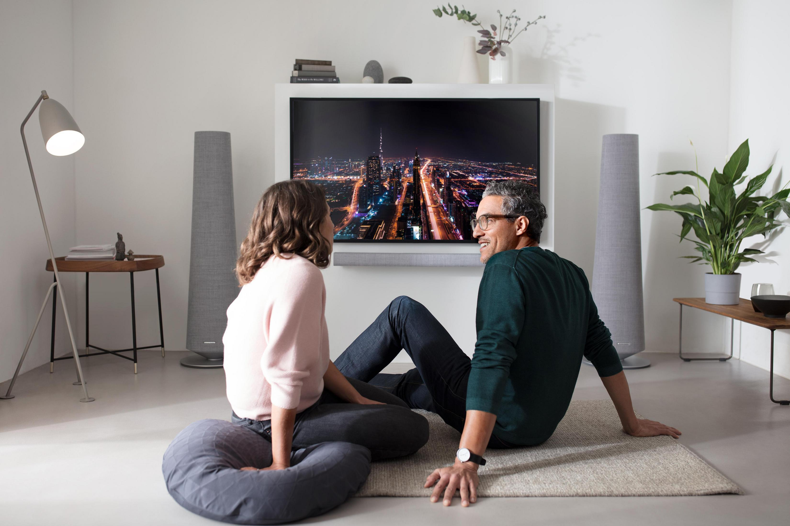 Ein Paar sitzt auf einem Teppich und schaut fern. Hohe Lautsprecher flankieren den Fernseher.