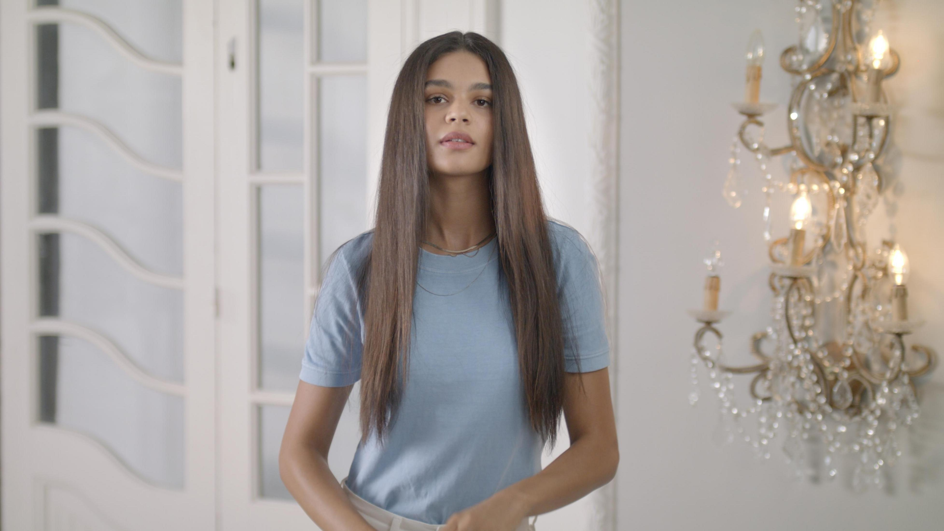 Vrouw in een blauw shirt. Staat in een kamer met een kroonluchter en witte deuren.