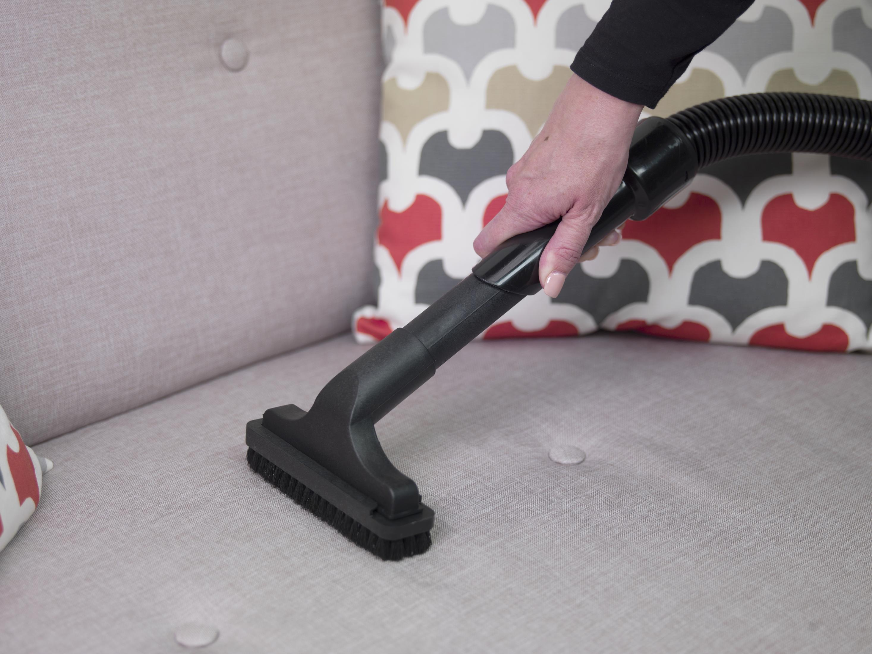 Ręka osoby odkurza szarą sofę czarną głowicą odkurzacza, z wzorzystymi poduszkami.