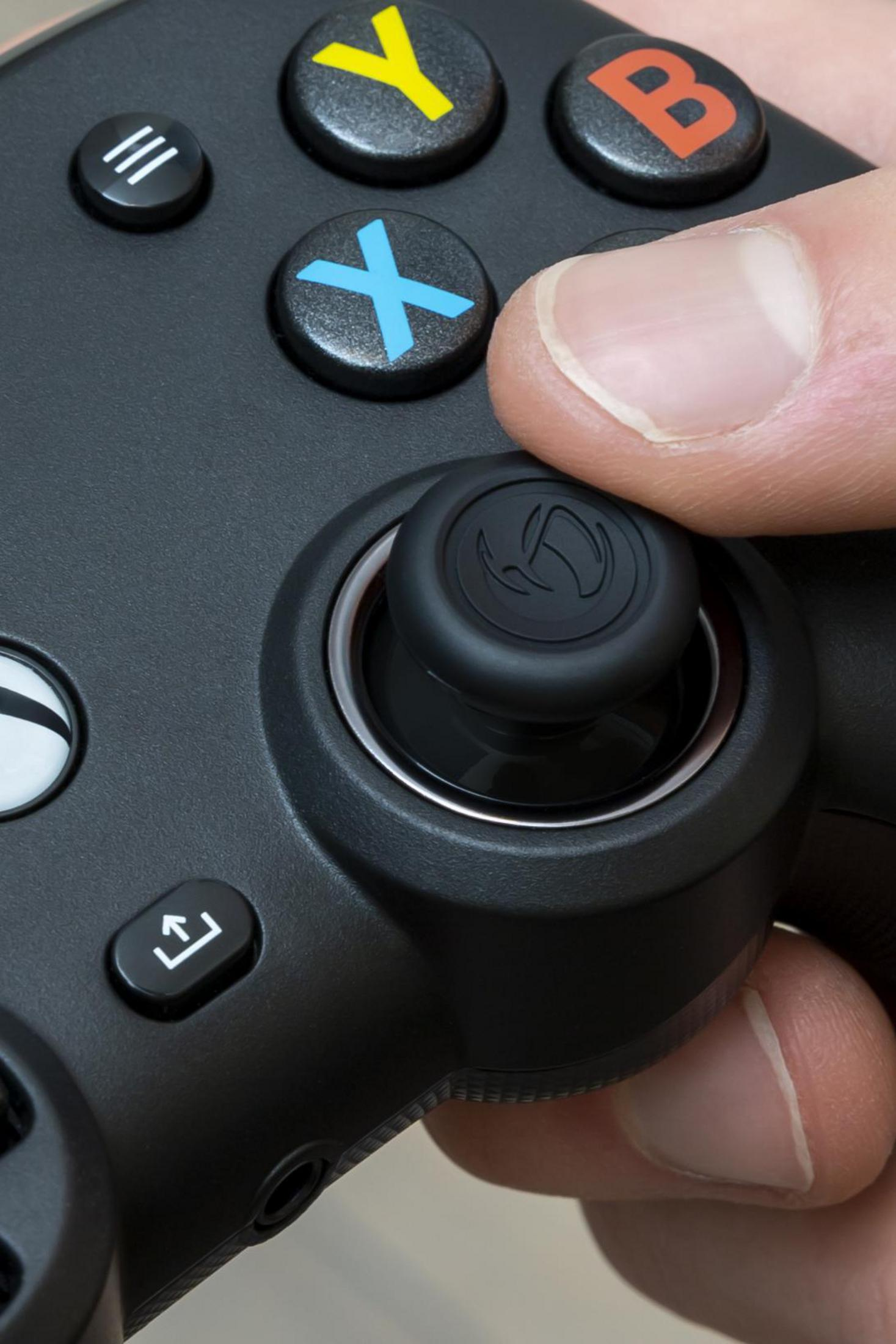 Dłoń trzymająca czarny gamepad. Kciuk jest na joysticku. Przyciski są widoczne.