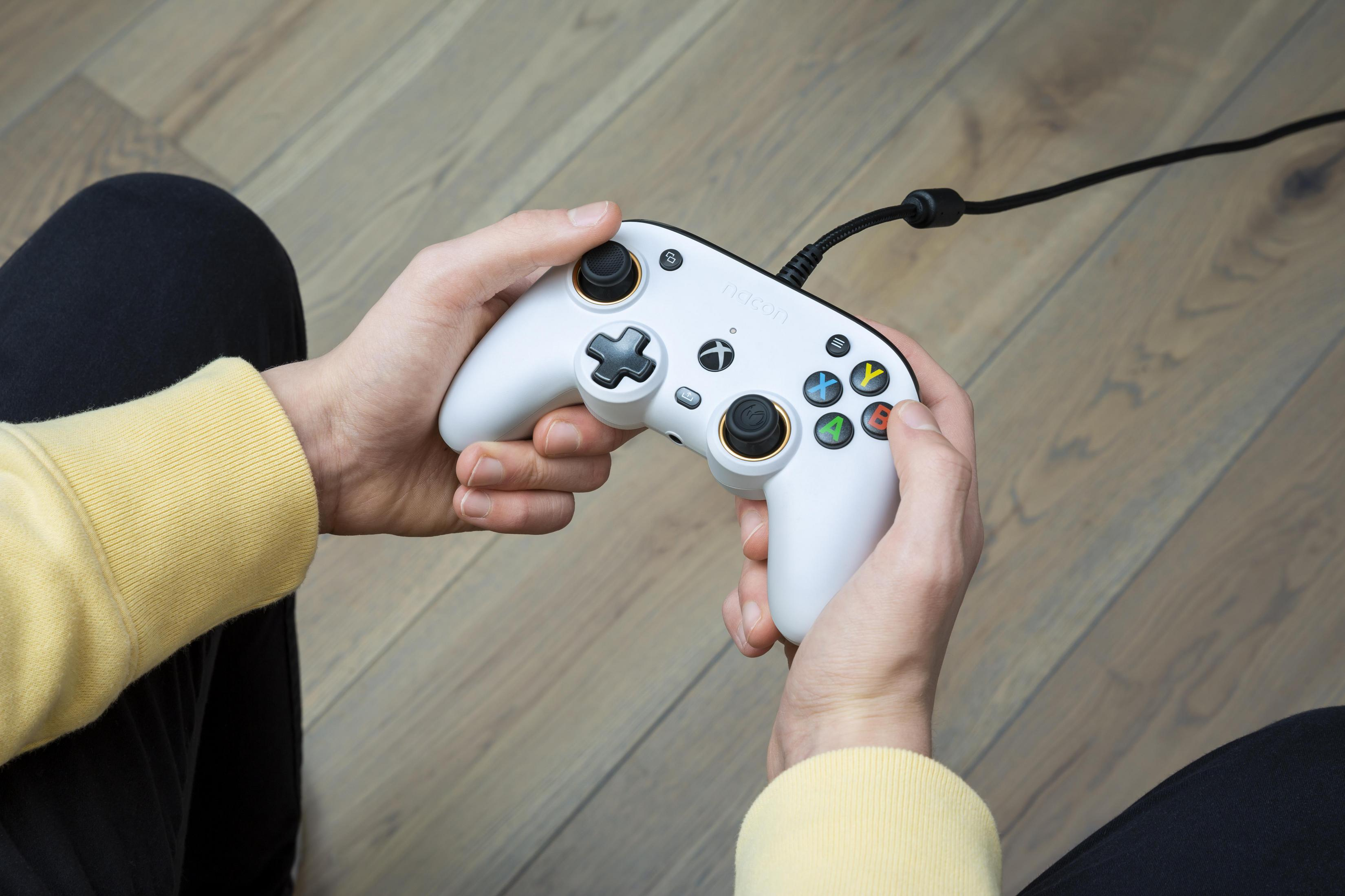 Biały kontroler Xbox trzymany w dłoniach. Podłączony czarny kabel. Widoczna żółta koszulka z długim rękawem.