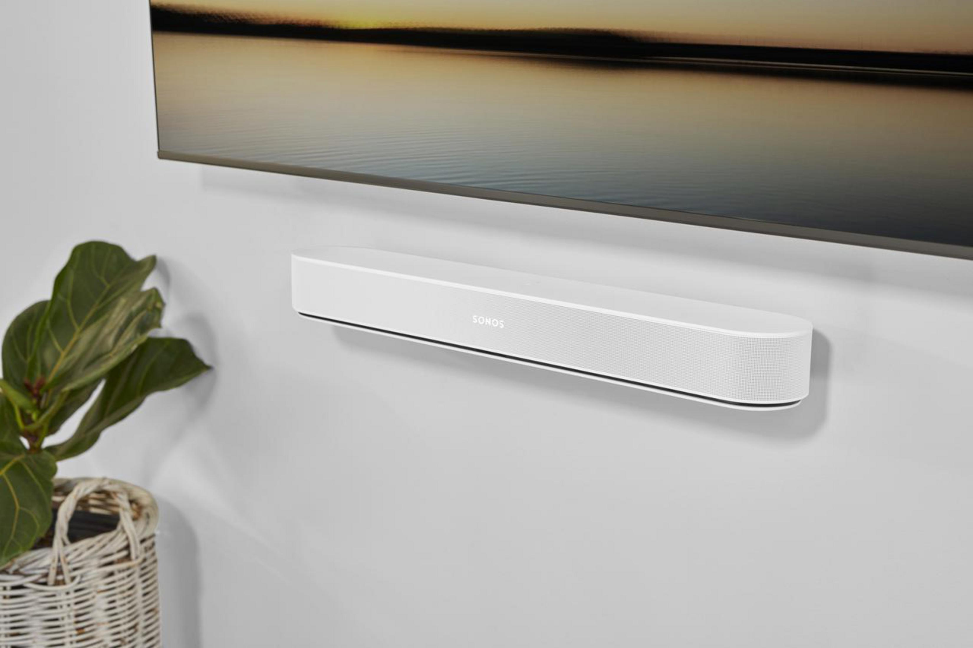 Soundbar bianca montata su una parete bianca sotto una televisione. Pianta e cestino.