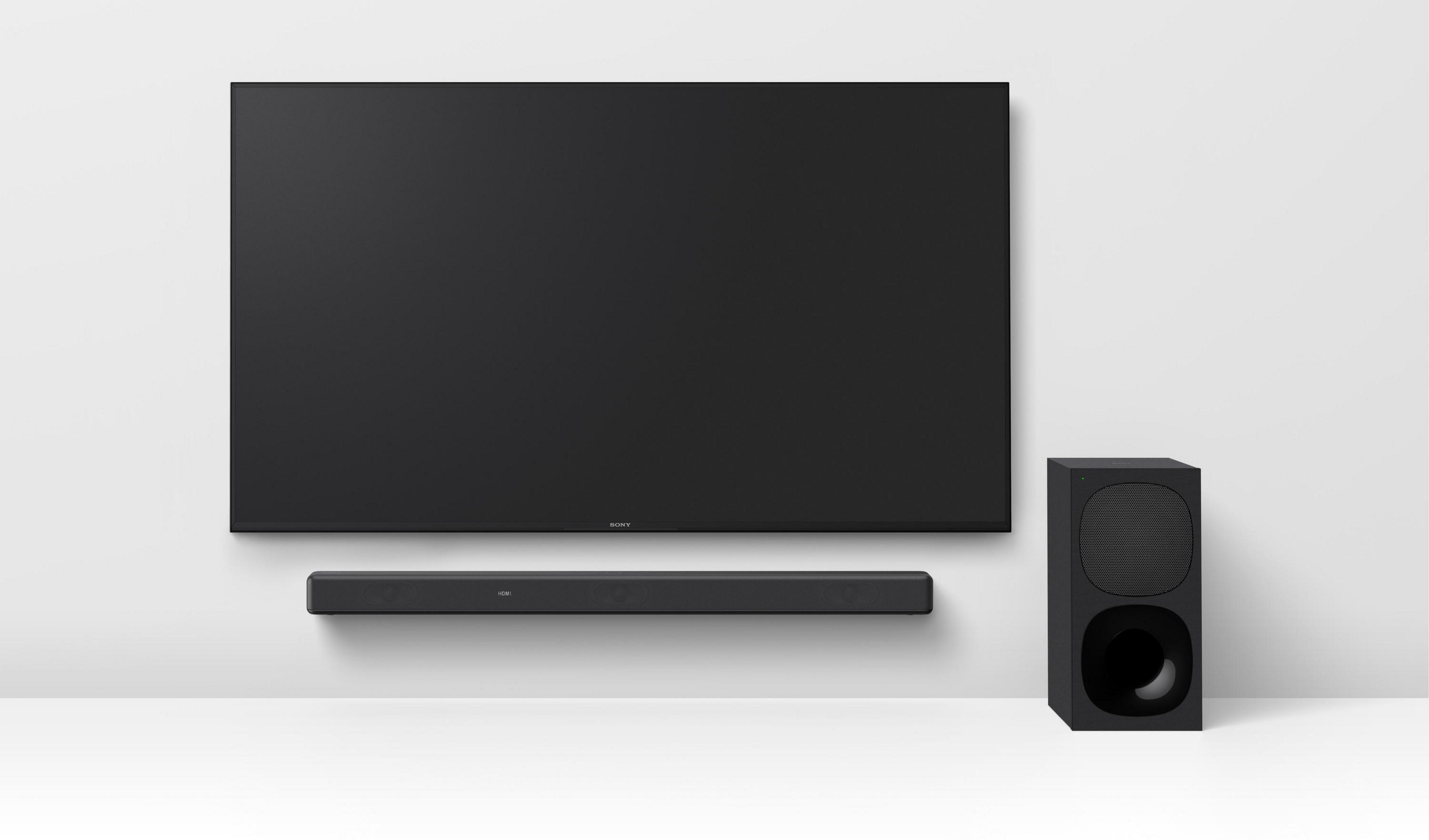 TV nero a parete sopra soundbar e subwoofer. Parete bianca, dispositivi grigio scuro. Estetica semplice e moderna.