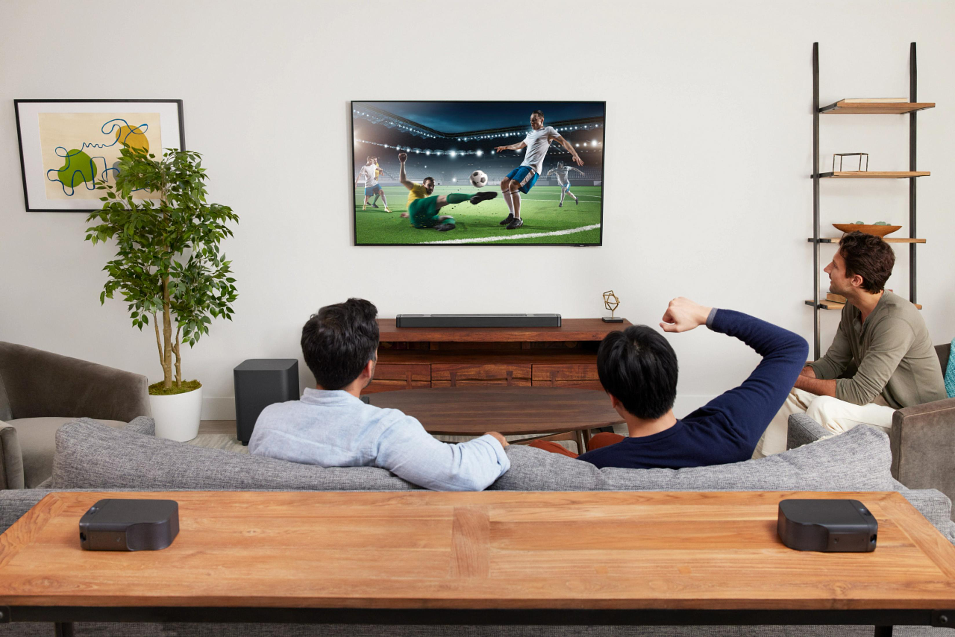 Persone guardano la TV con una soundbar. Due altoparlanti. Tavolo di legno.