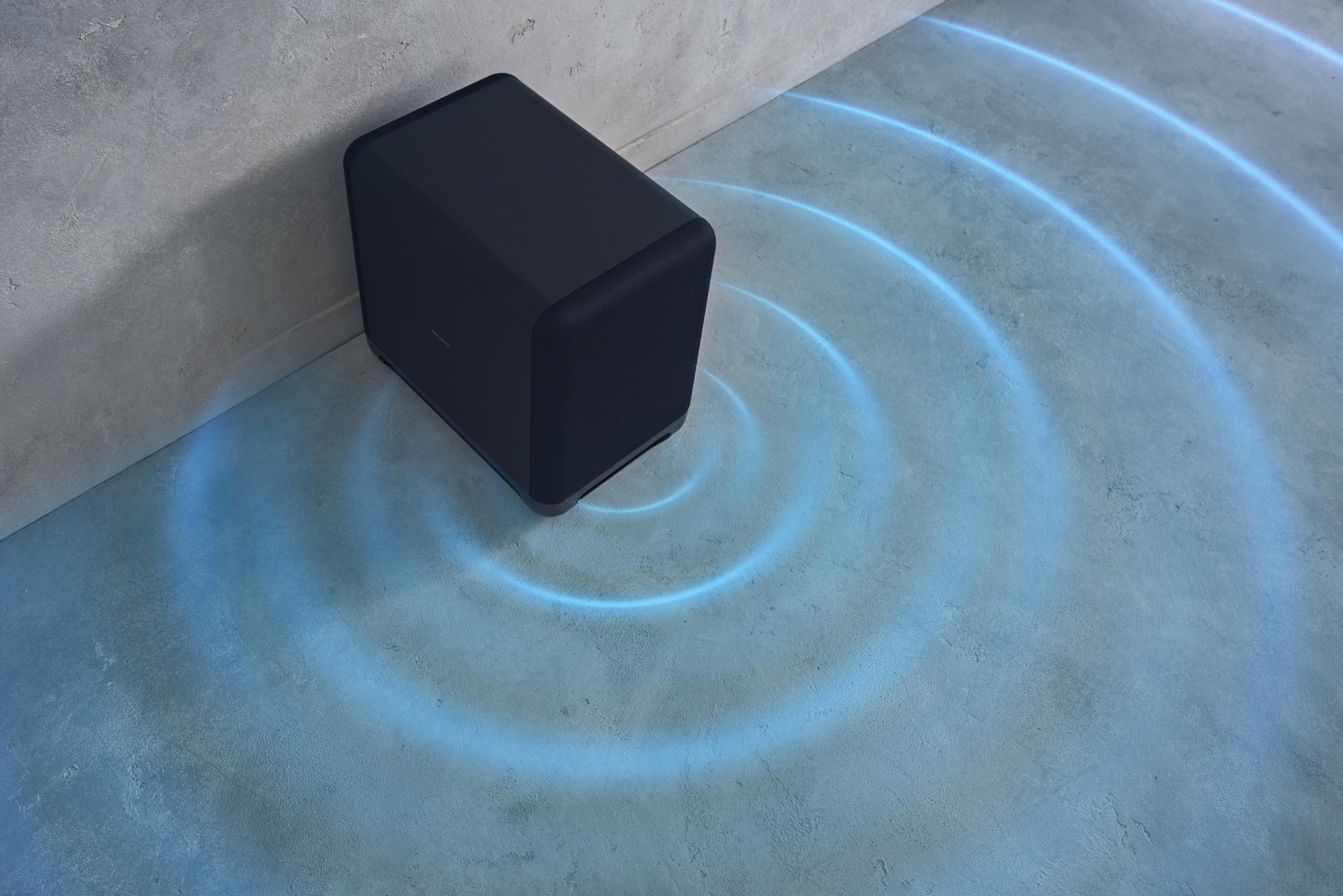 Czarny subwoofer na betonowej podłodze, z efektem niebieskiej fali dźwiękowej. Oparty o szarą ścianę.