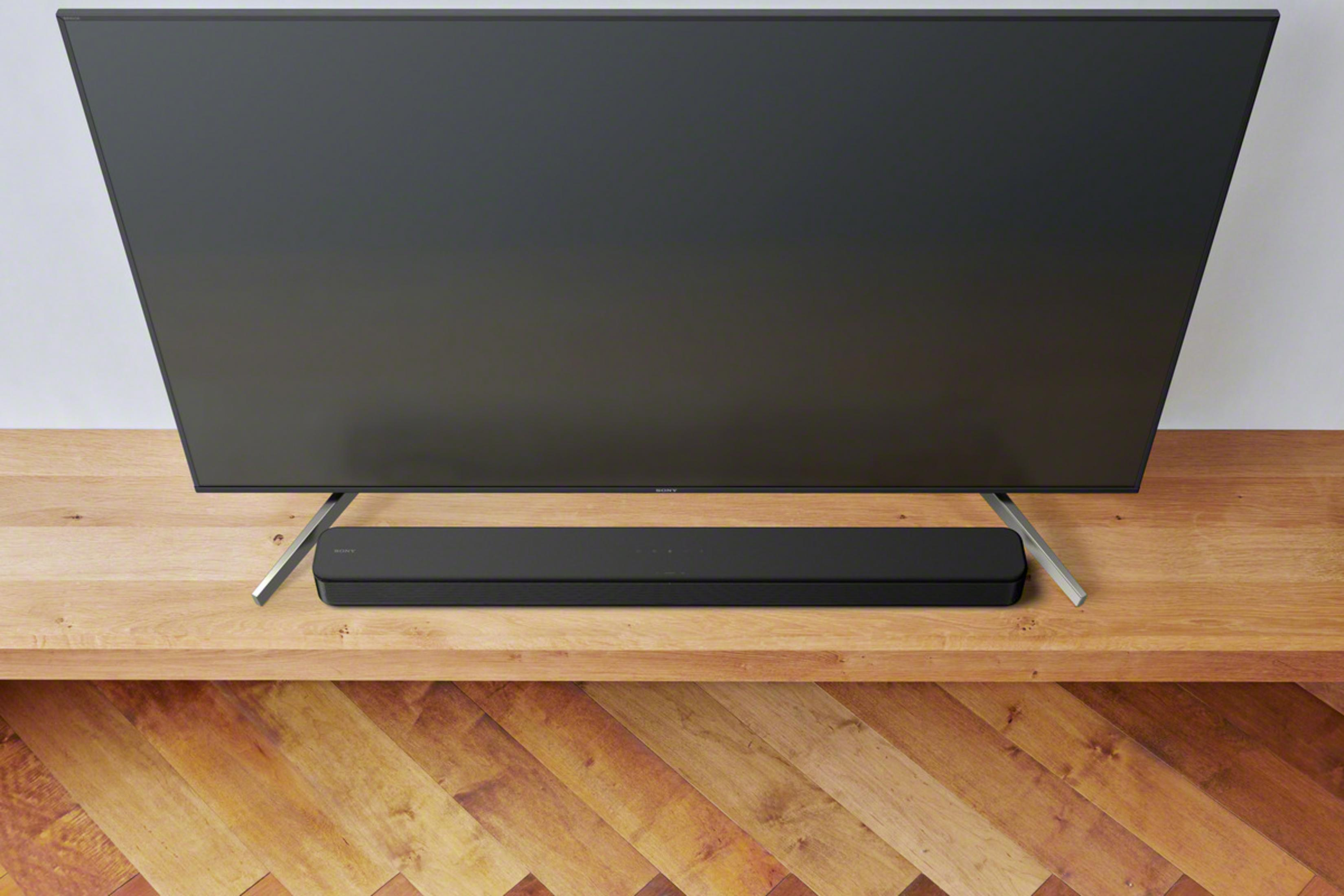 Ein großer schwarzer Fernseher über einer schwarzen Soundbar auf einer Holzoberfläche vor weißer Wand.