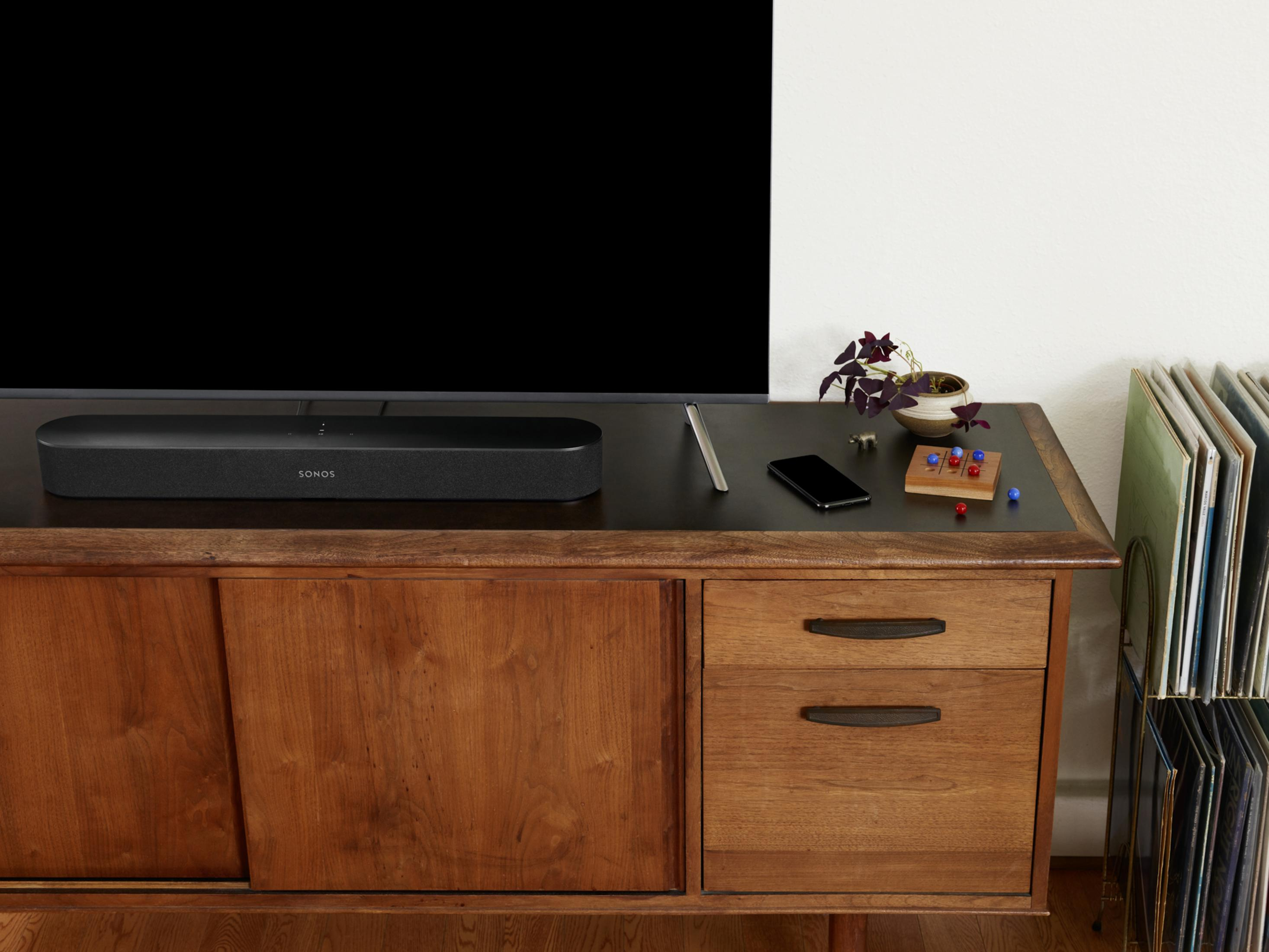 Una barra de sonido negra en un mueble de madera, un televisor encima. Varios objetos en el mueble.