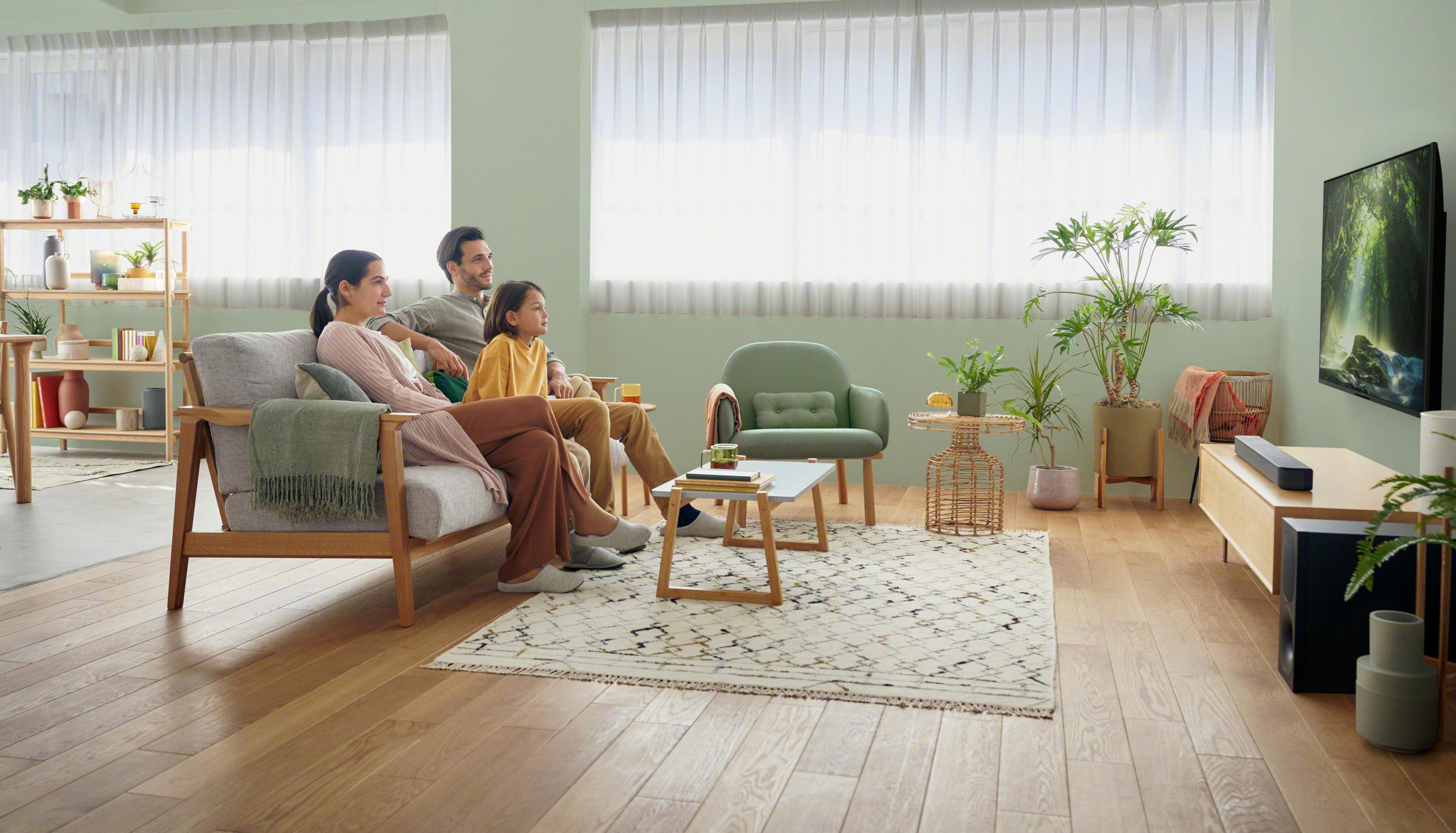 Familie schaut im Wohnzimmer fern. Sofa, Teppich und Pflanzen sind sichtbar.