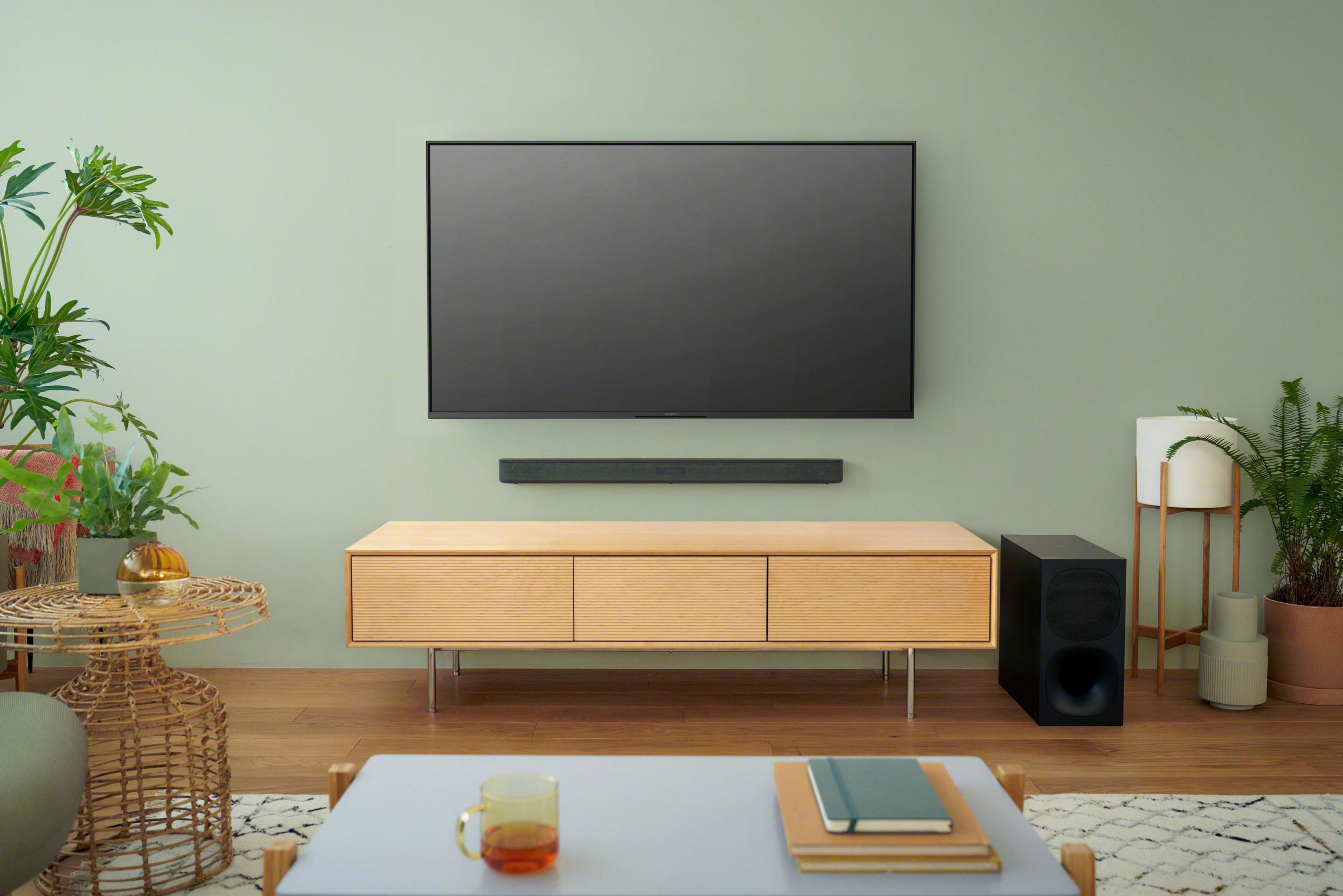 Wohnzimmer mit TV, Soundbar und Holzschrank. Grüne Wand, Pflanzen.