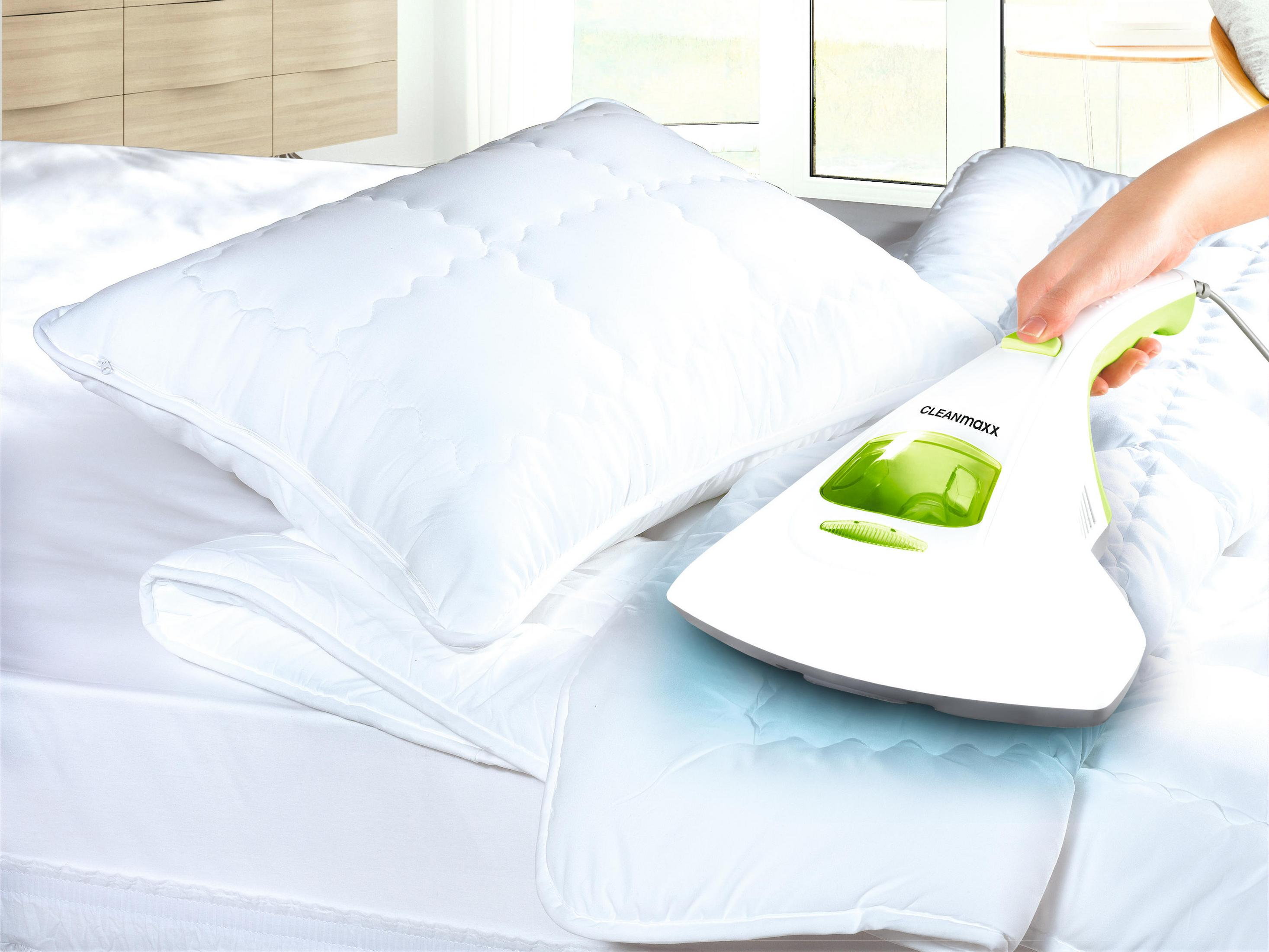 Ręka trzyma biało-zielony odkurzacz nad białą poduszką i kocem na łóżku.