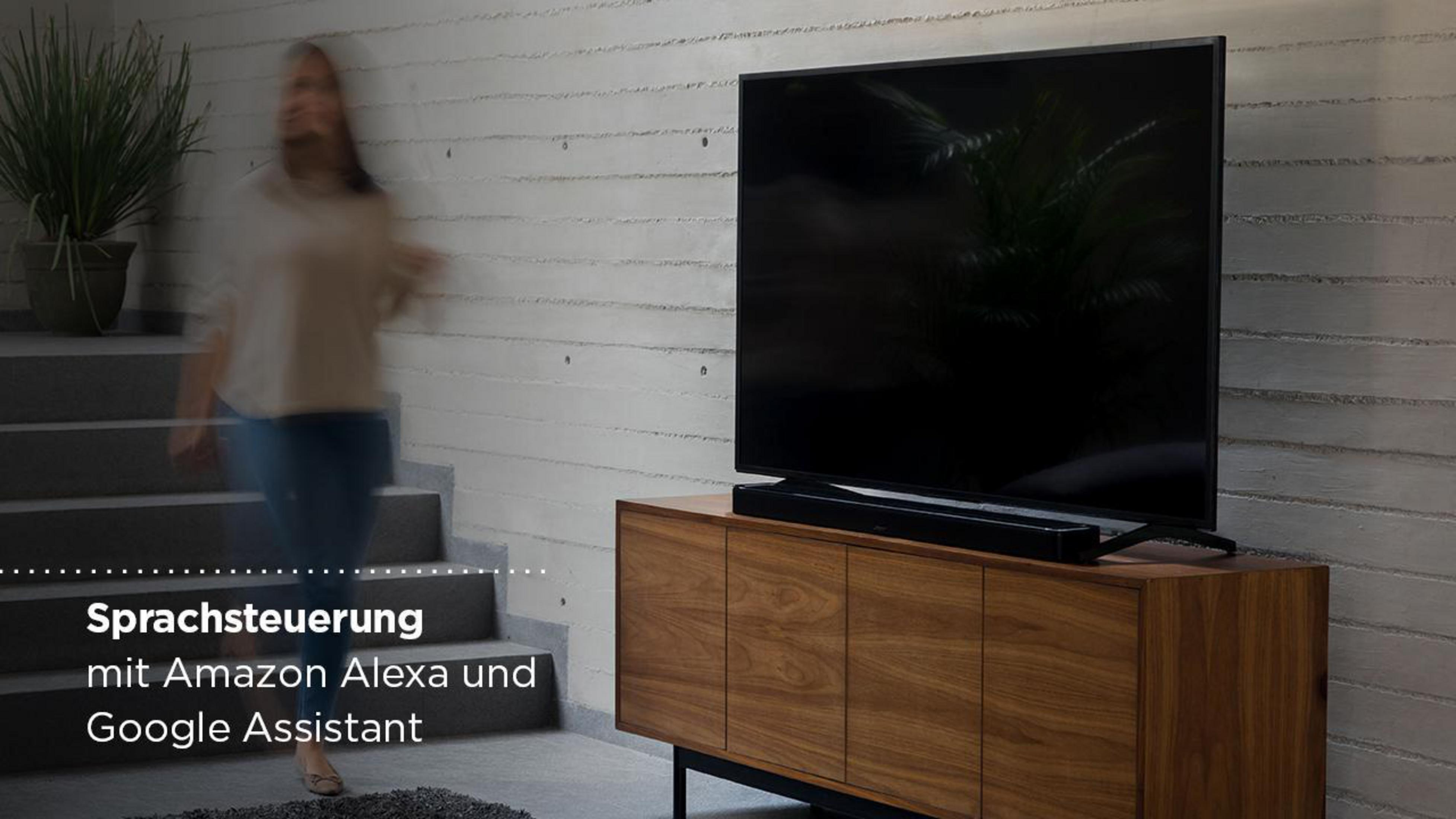 Ein Fernseher auf einem Holzschrank. Eine Frau geht davor. Text sagt Sprachsteuerung mit Amazon Alexa und Google Assistant.