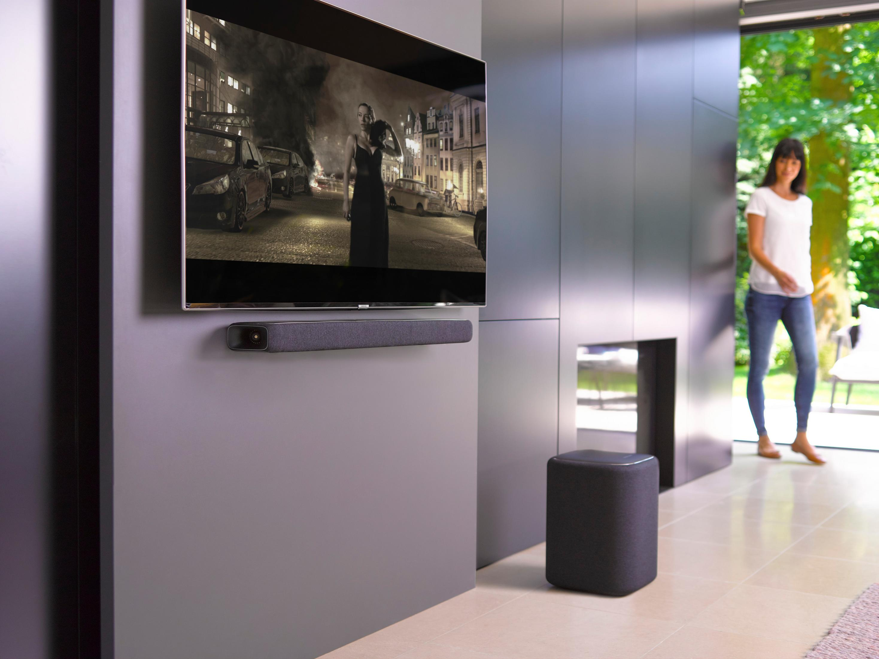 Eine Frau geht auf einen Fernseher und eine Soundbar zu, ein Lautsprecher steht auf dem Boden. Der Fernseher zeigt einen Film.