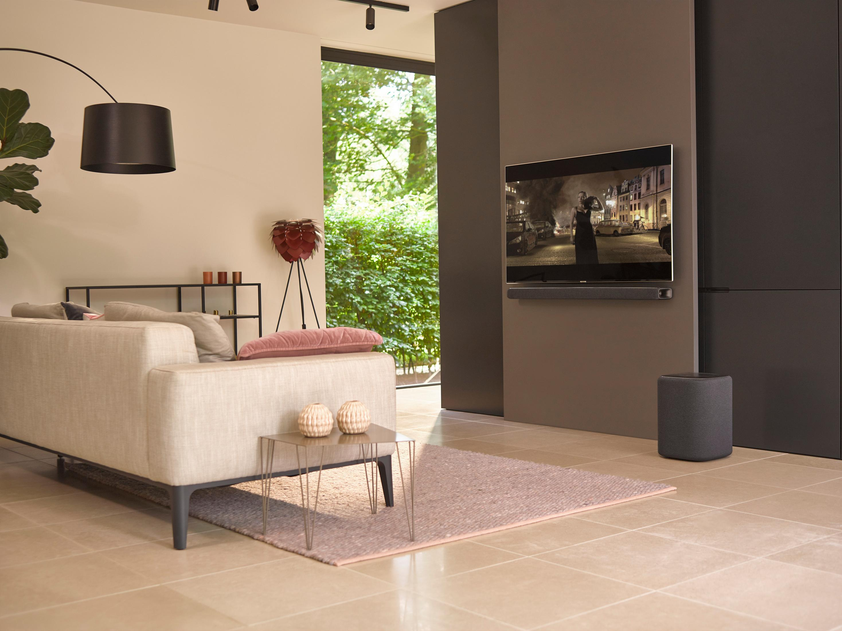 Wohnzimmer mit Sofa, Couchtisch und Fernseher. Der Fernseher zeigt einen Film. Ein Subwoofer steht auf dem Boden.