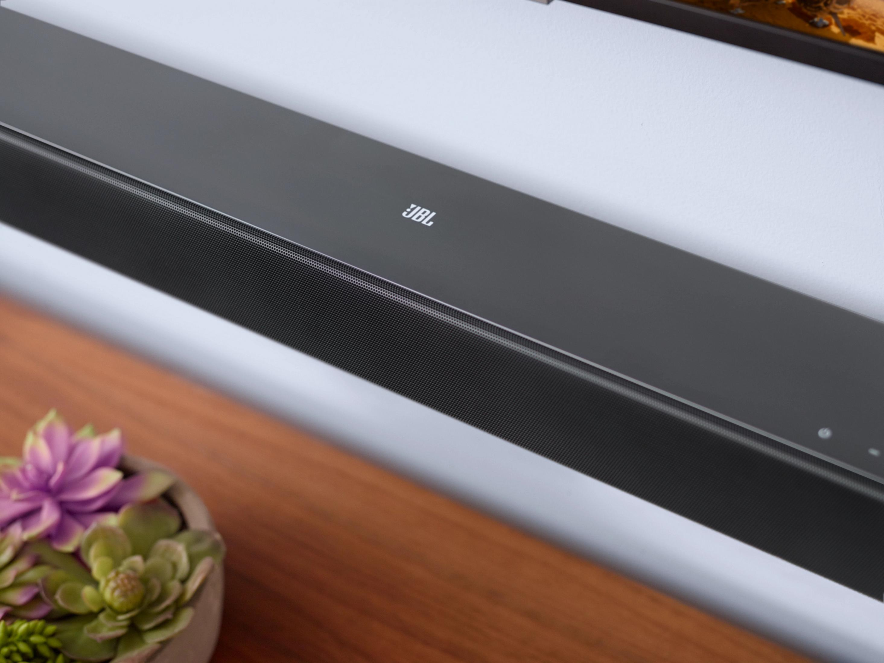 Schwarze Soundbar an einer weißen Wand, mit einer kleinen Pflanze im Vordergrund.