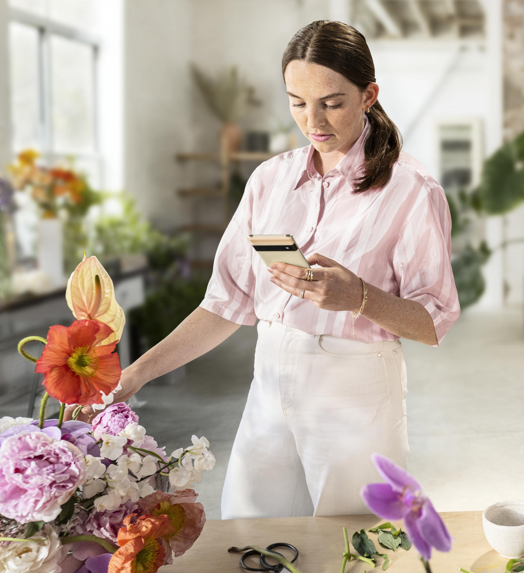 Kobieta w różowej koszuli z telefonem przy kwiatach. Kwiaty to pomarańczowe maki i różowe piwonie. Rozmyte tło.