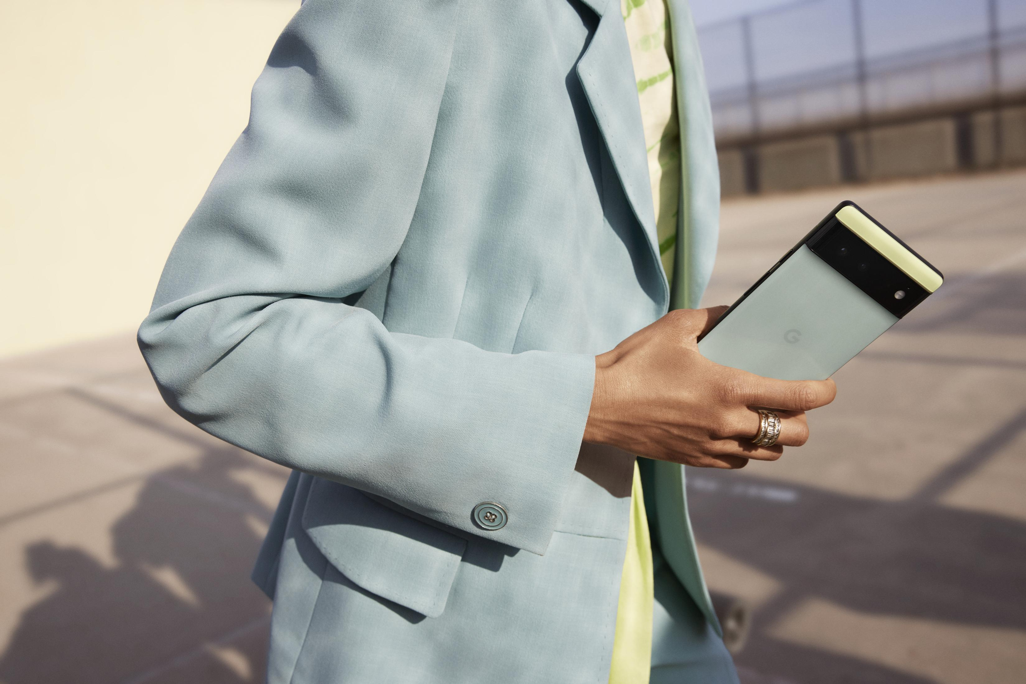 Eine Person hält ein hellgrünes Pixel 6a Telefon. Trägt einen hellblauen Anzug.