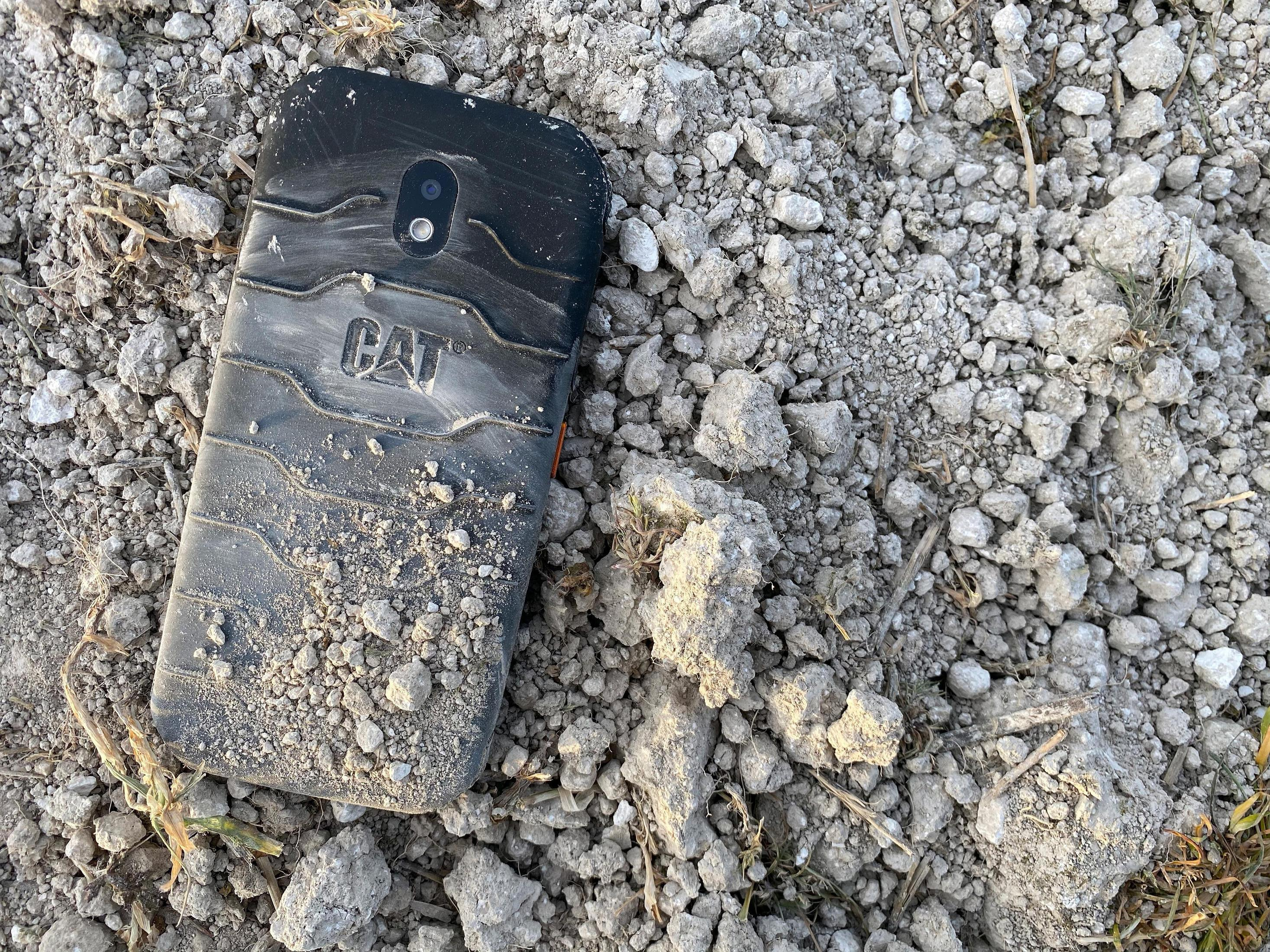 Ein schwarzes CAT-Telefon, das mit Schmutz bedeckt ist, liegt auf einem Boden aus kleinen Steinen und Erde.