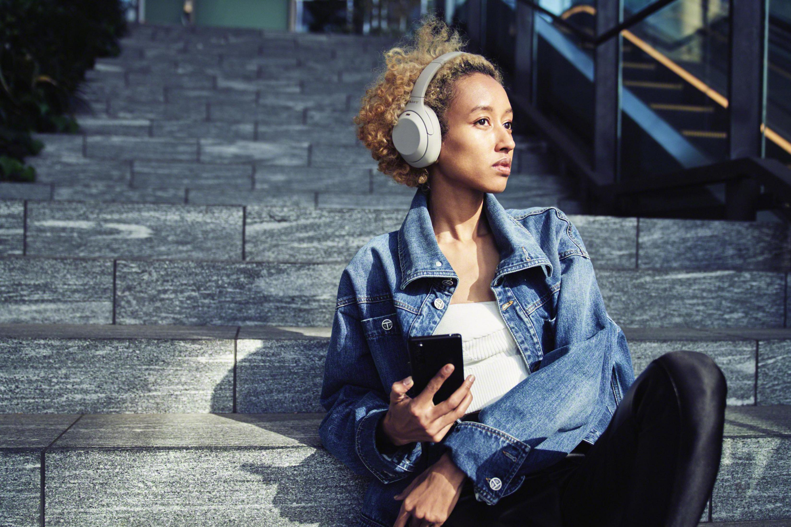 Kobieta ze słuchawkami i telefonem siedzi na schodach, patrząc w bok. Beton i metal.