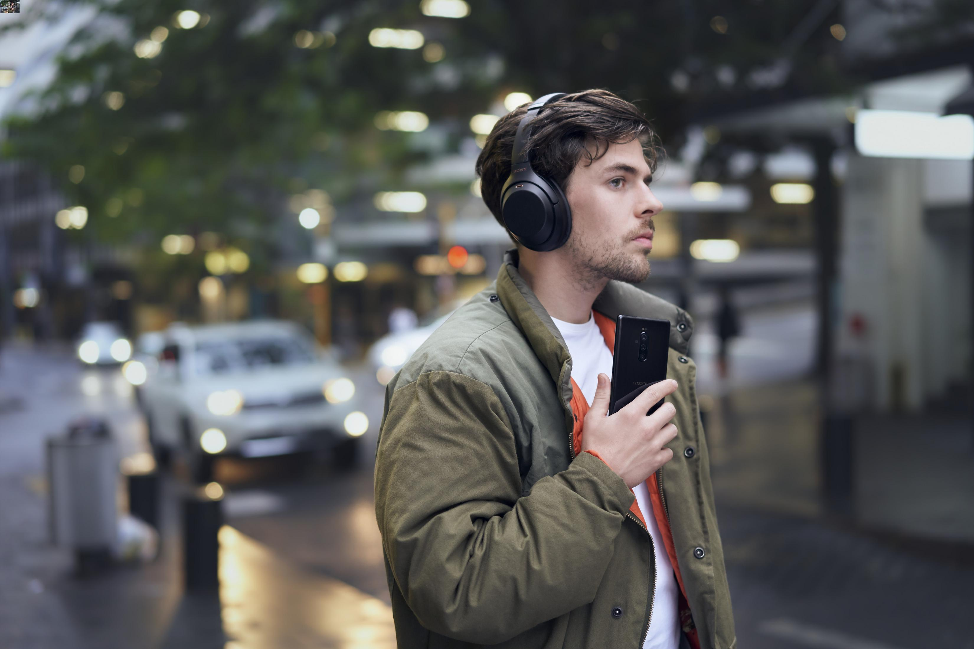 Mann mit Kopfhörern, hält ein Smartphone, geht auf einer Stadtstraße.