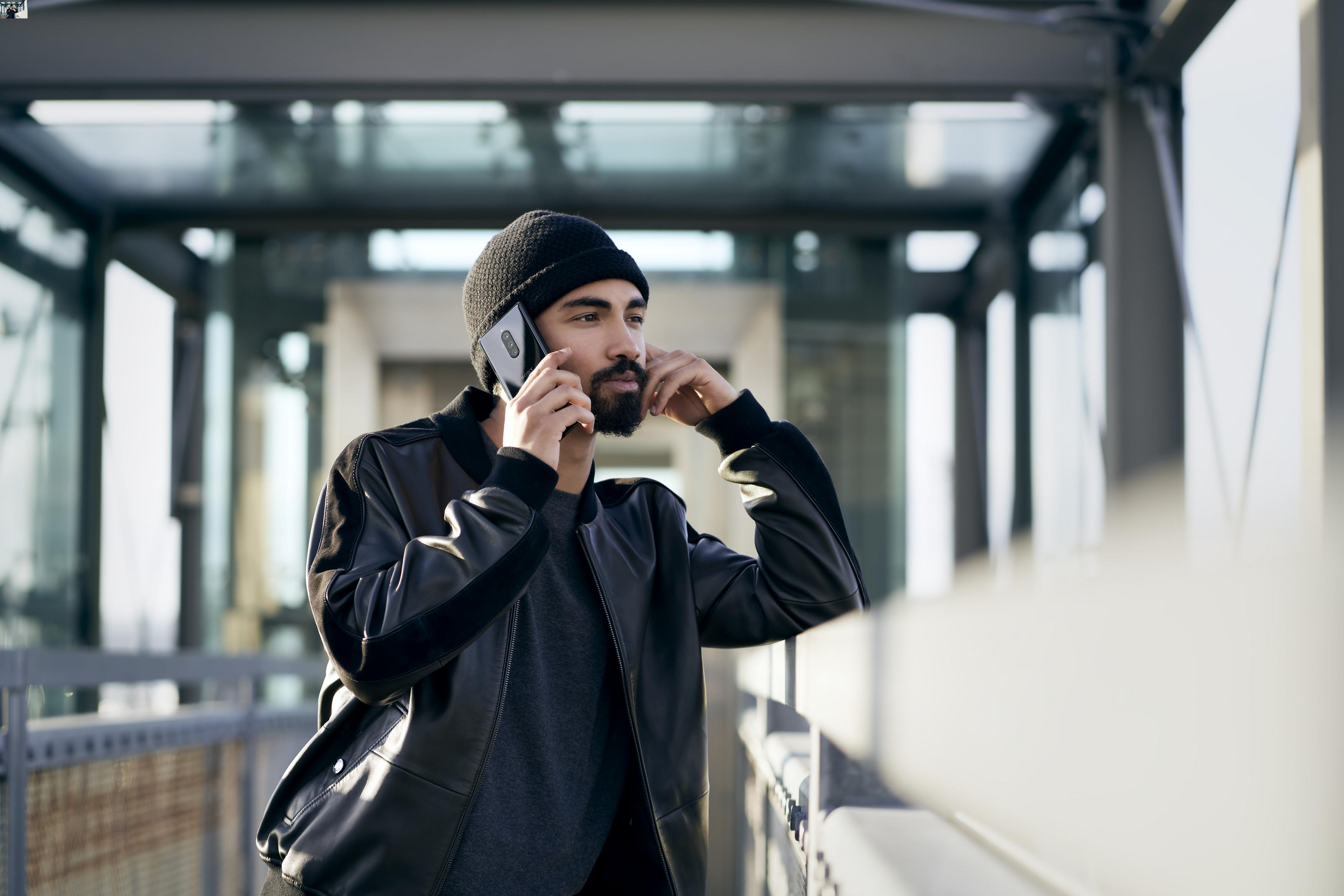 Ein Mann mit schwarzer Mütze und Jacke telefoniert. Er steht auf einer Brücke.