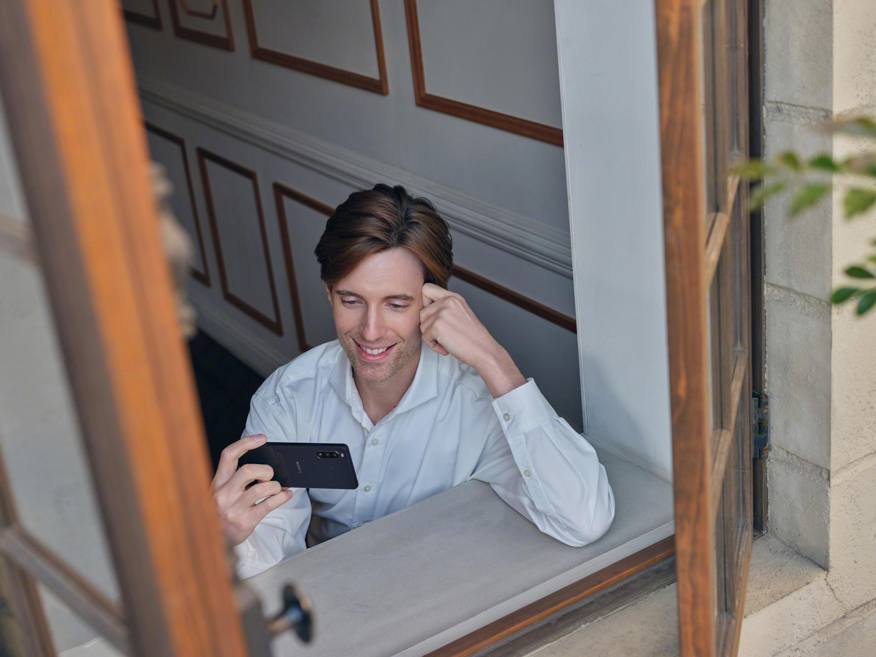 Man smiling while looking at a phone, sitting in a window frame.
