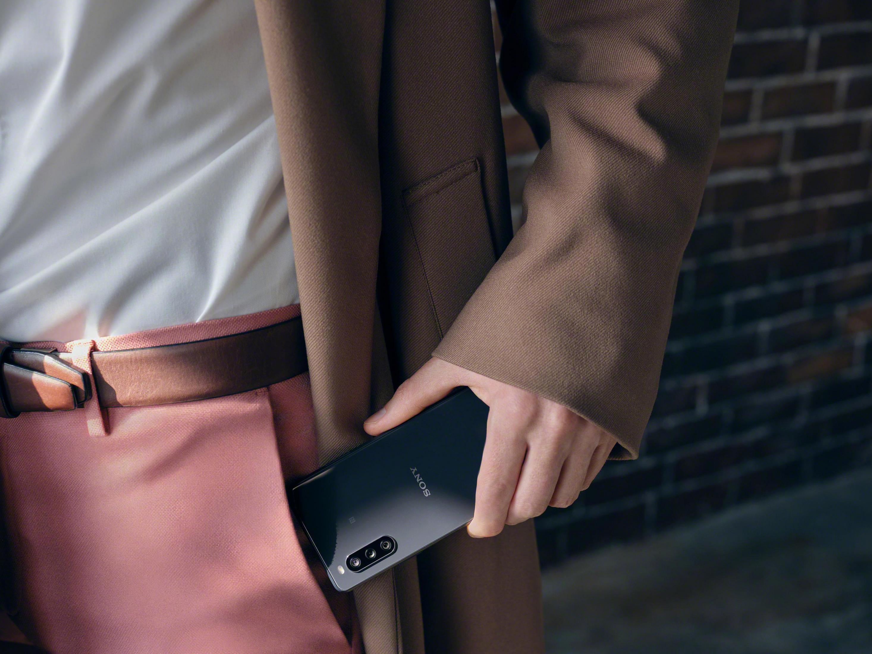 A hand holds a black Sony phone in a pocket. Background: pink pants, white shirt, and brown coat.