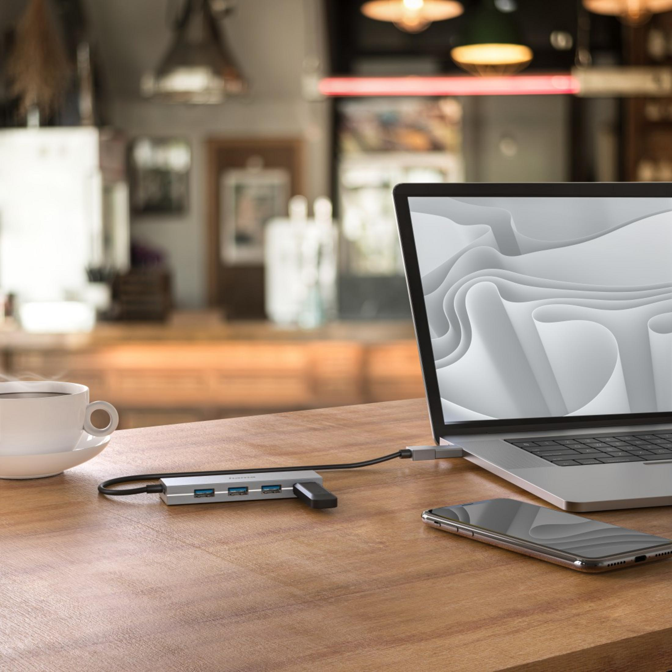 Un laptop con un hub e un telefono su un tavolo di legno. L'hub ha porte USB e un lettore di schede. È visibile anche una tazza di caffè.